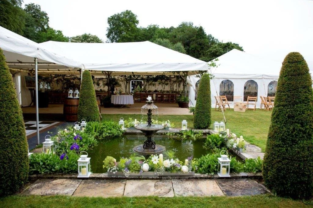 A decorated outdoor garden area with a small pond, a fountain, and white lanterns surrounded by flowers. There are large tents with seating and a waterfall on the edge of the pond.