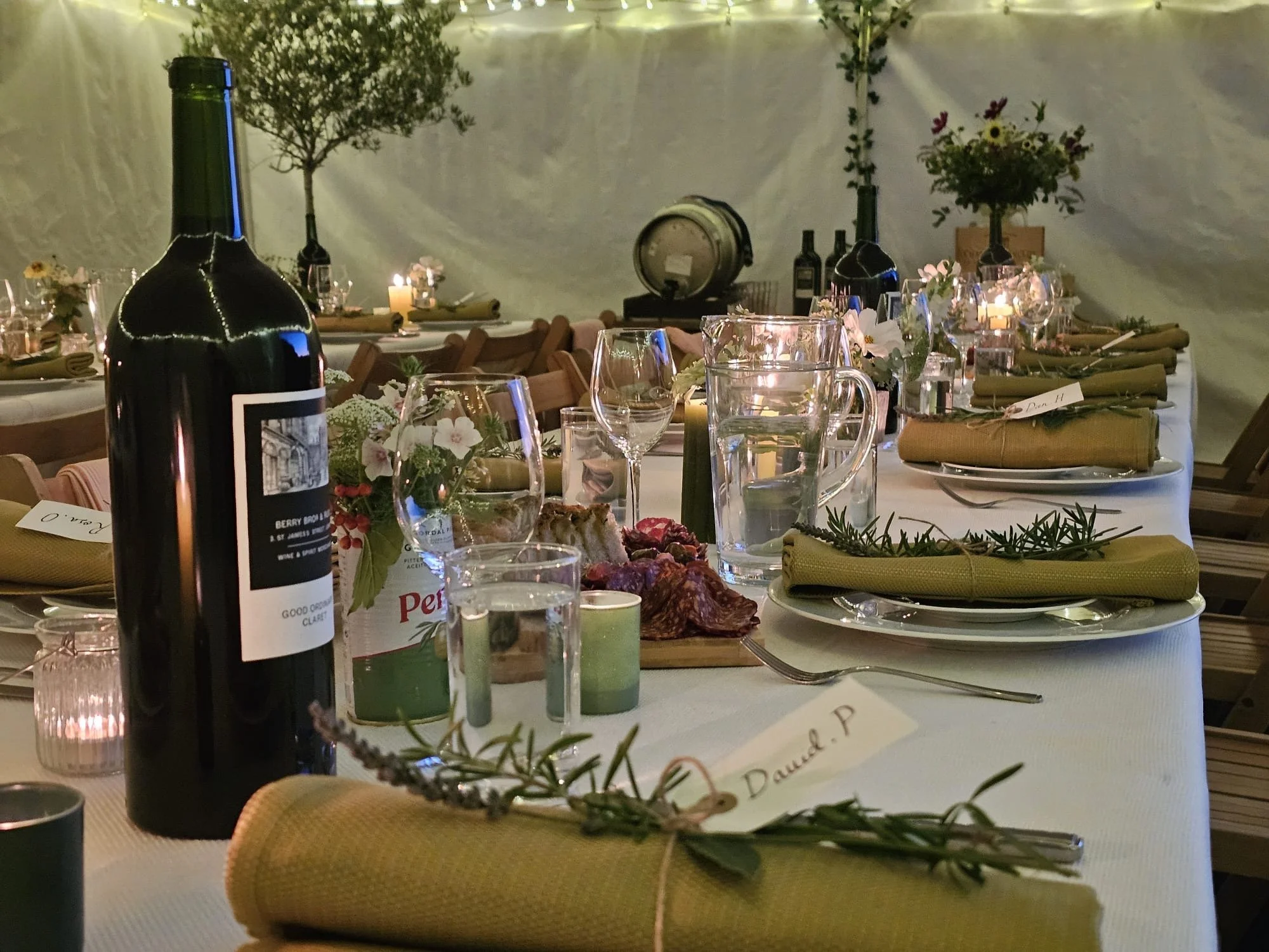 A banquet table in a decorated event tent with candles, wine bottles, flowers, and neatly rolled napkins, ready for a gathering or celebration.