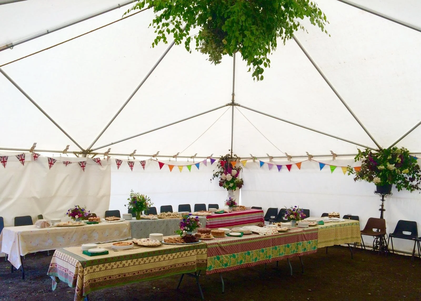 A decorated event tent with a long buffet table covered with colorful tablecloths, flowers, and baked goods, with bunting flags hanging along the interior ceiling.