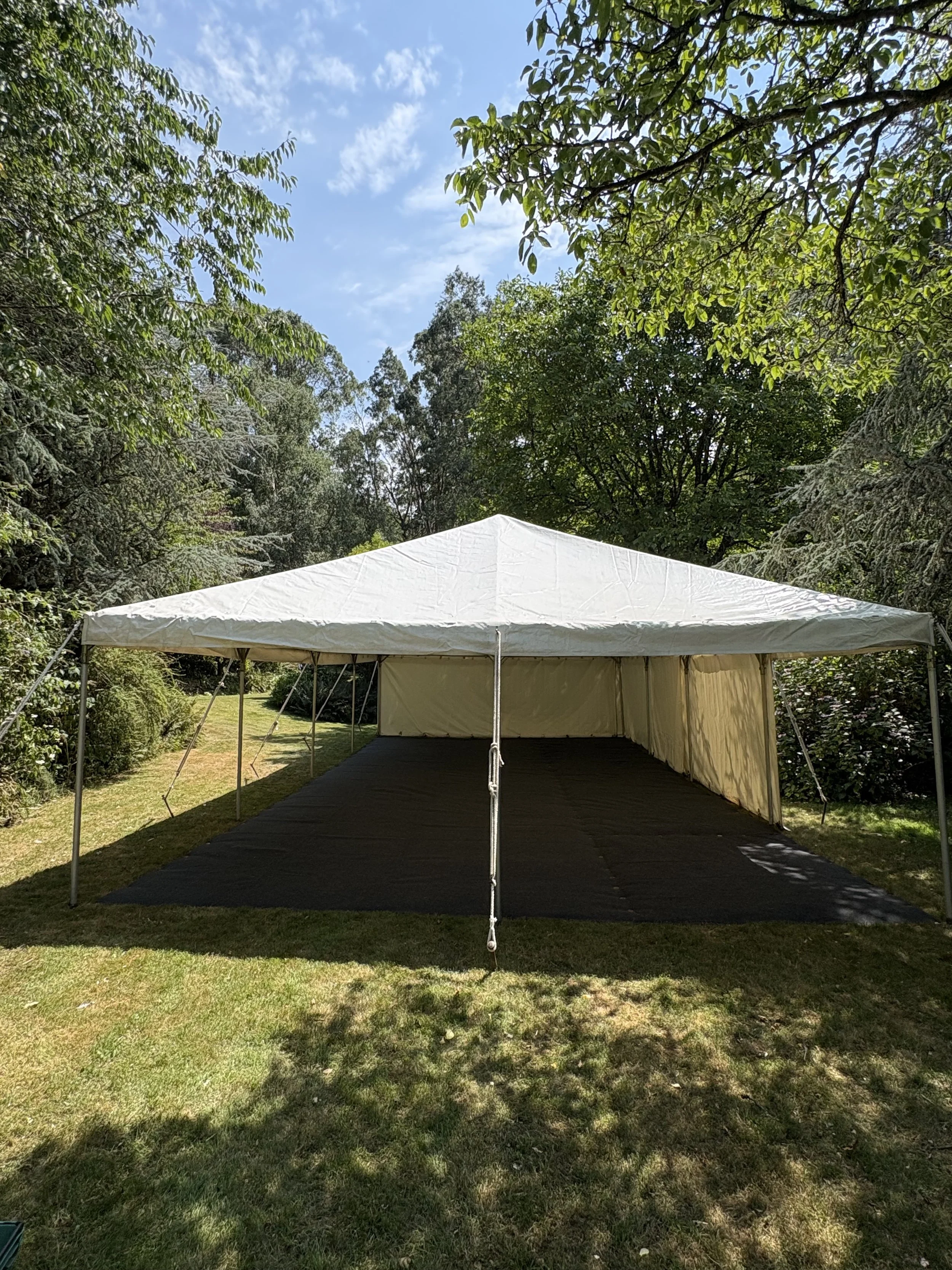 An outdoor white event tent set up on a grassy area surrounded by trees, with a partly cloudy sky above.