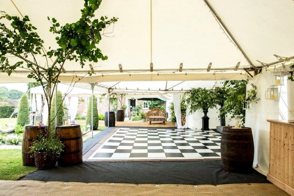 A large outdoor event tent with a checkered black and white dance floor, decorated with potted plants and barrels, with seating area in the background, set up for a celebration or gathering.