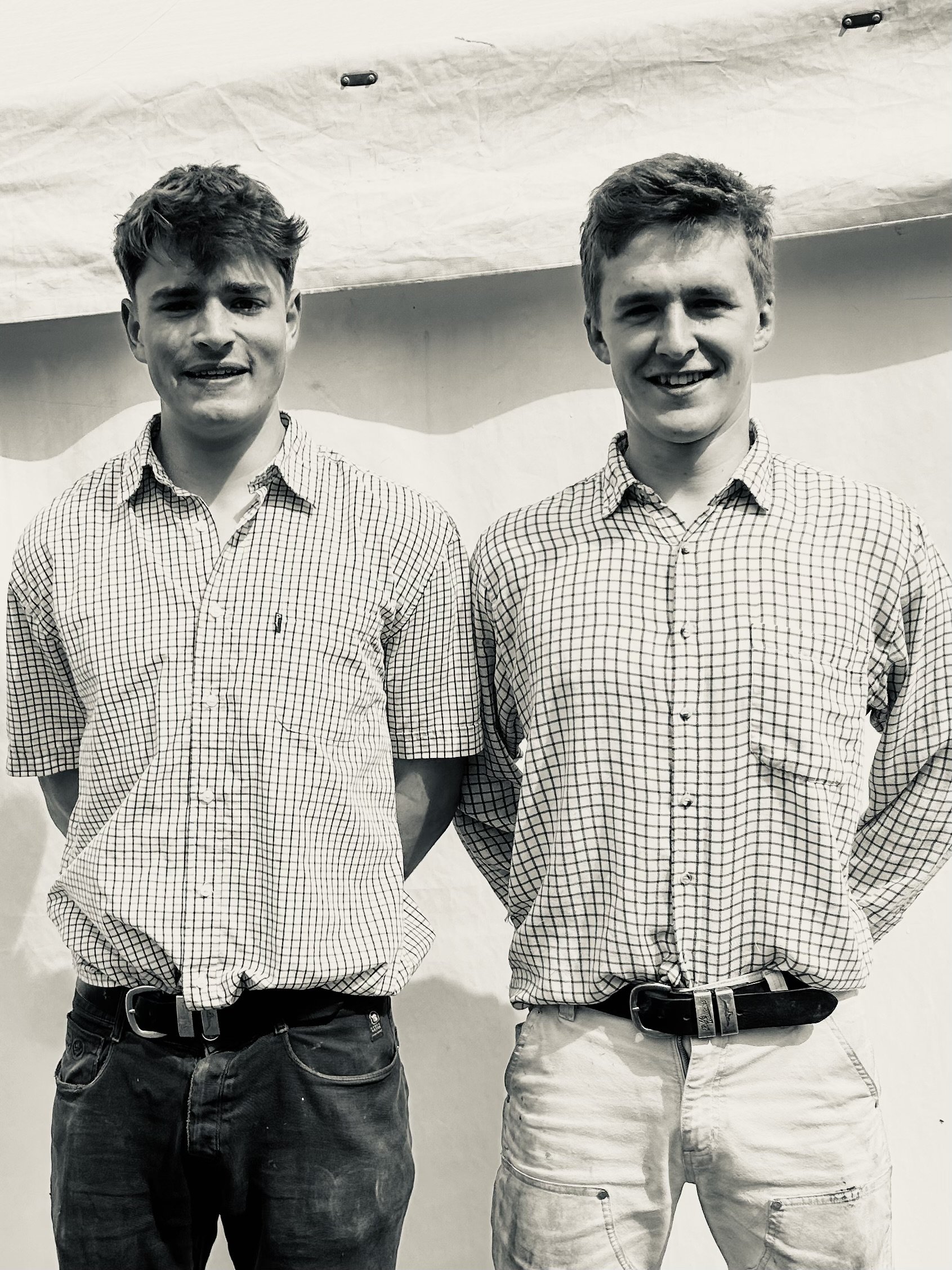 Two young men standing side by side, both wearing checkered shirts and jeans, smiling at the camera, in front of a plain backdrop.