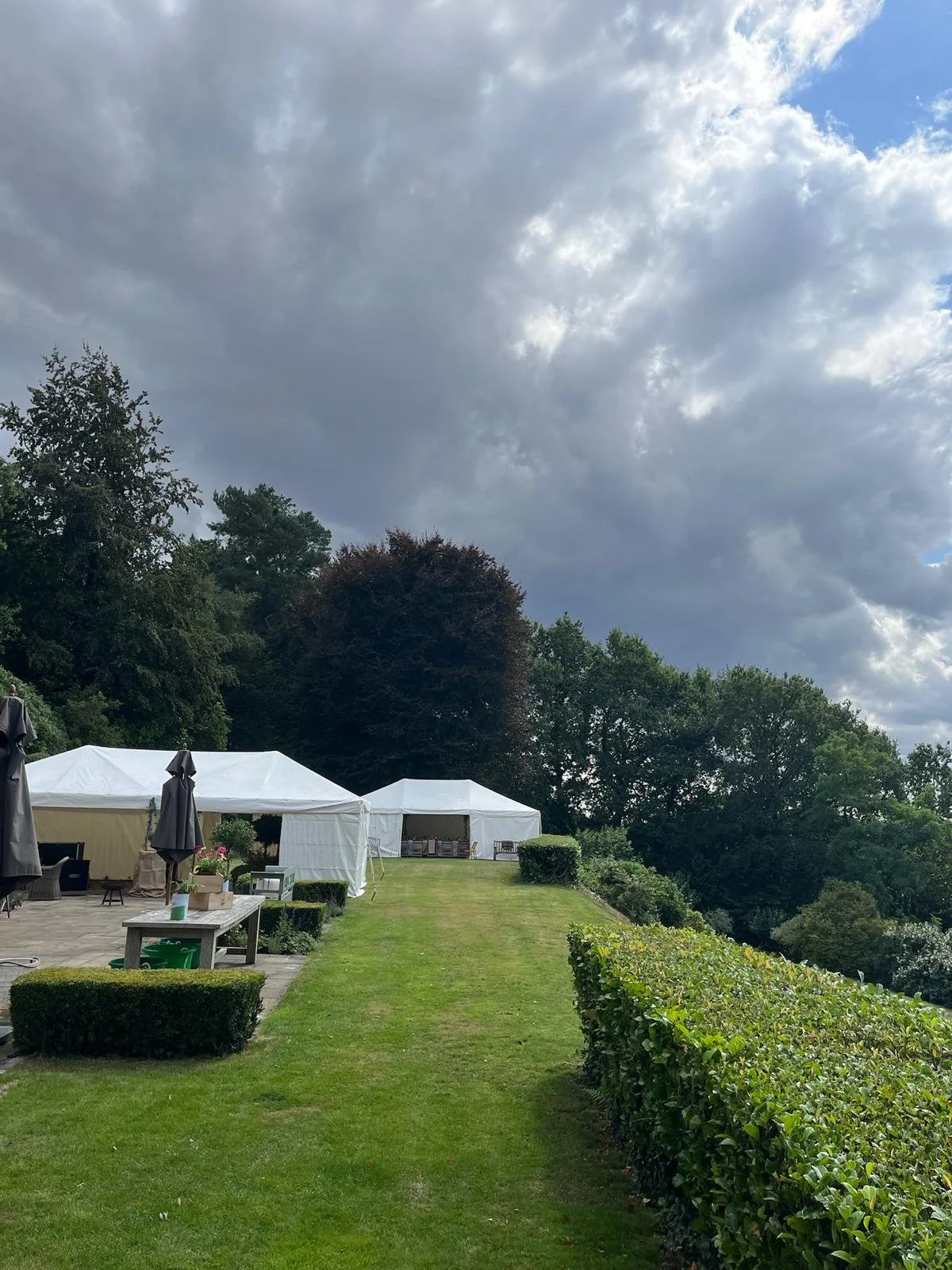 An outdoor backyard scene with white tents, a patio with umbrellas, a small table, and a well-maintained lawn surrounded by shrubs and trees under a cloudy sky.