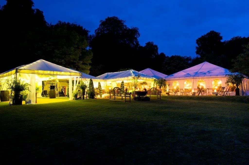 Outdoor event tents illuminated with lights at night, surrounded by trees and chairs on a grassy area.
