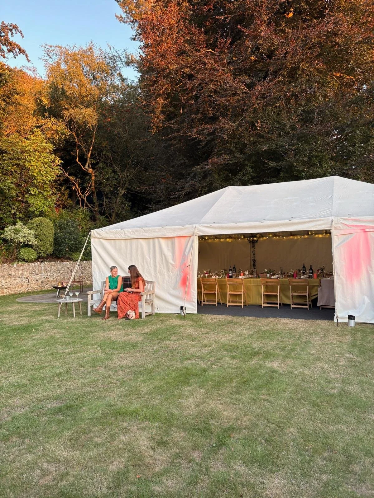 Two women sitting and talking outside a white event tent with a long table and chairs set up inside, on a grassy lawn with trees in the background.