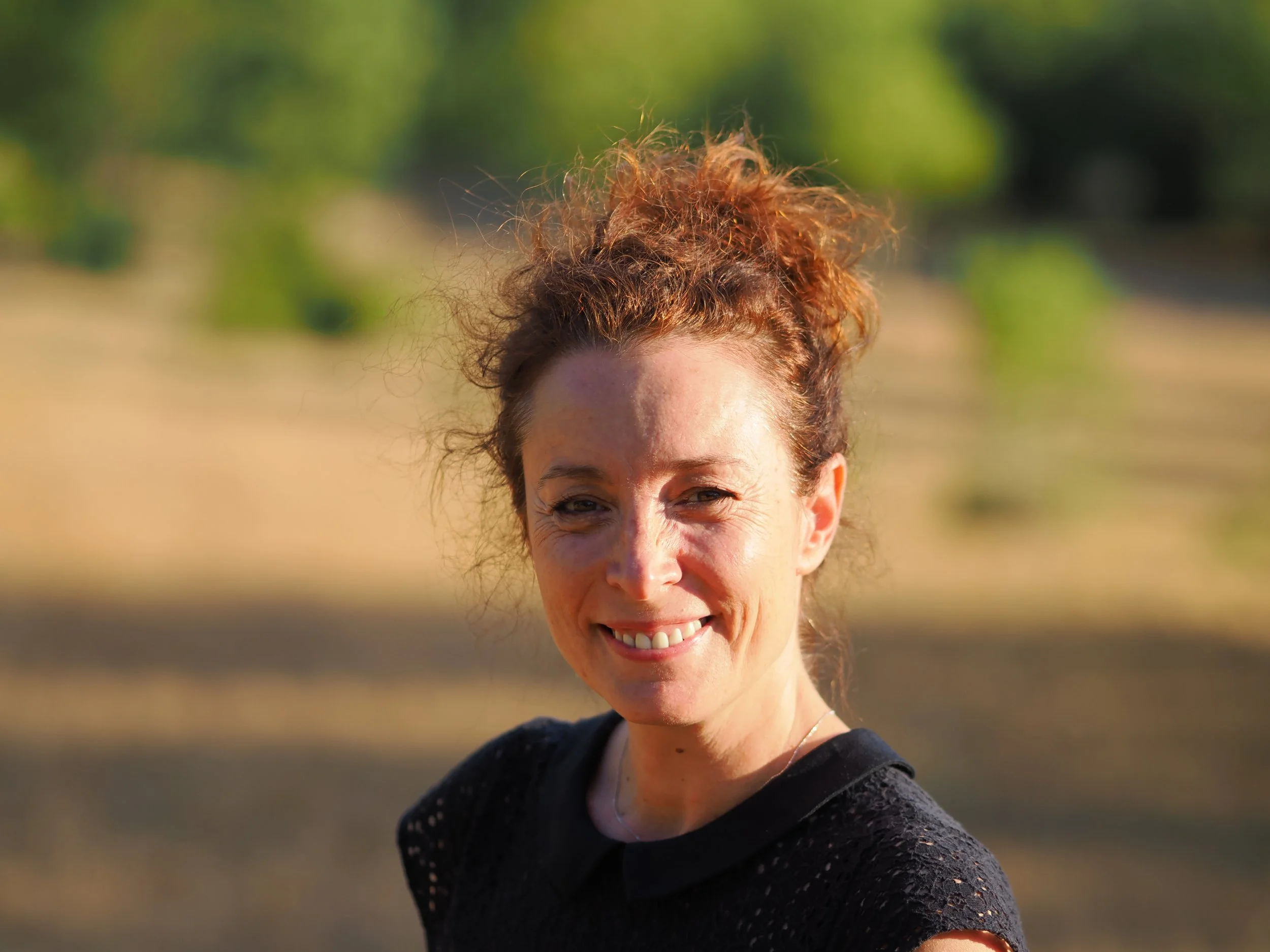 A woman with curly red hair tied in a high bun, smiling outdoors in natural light.