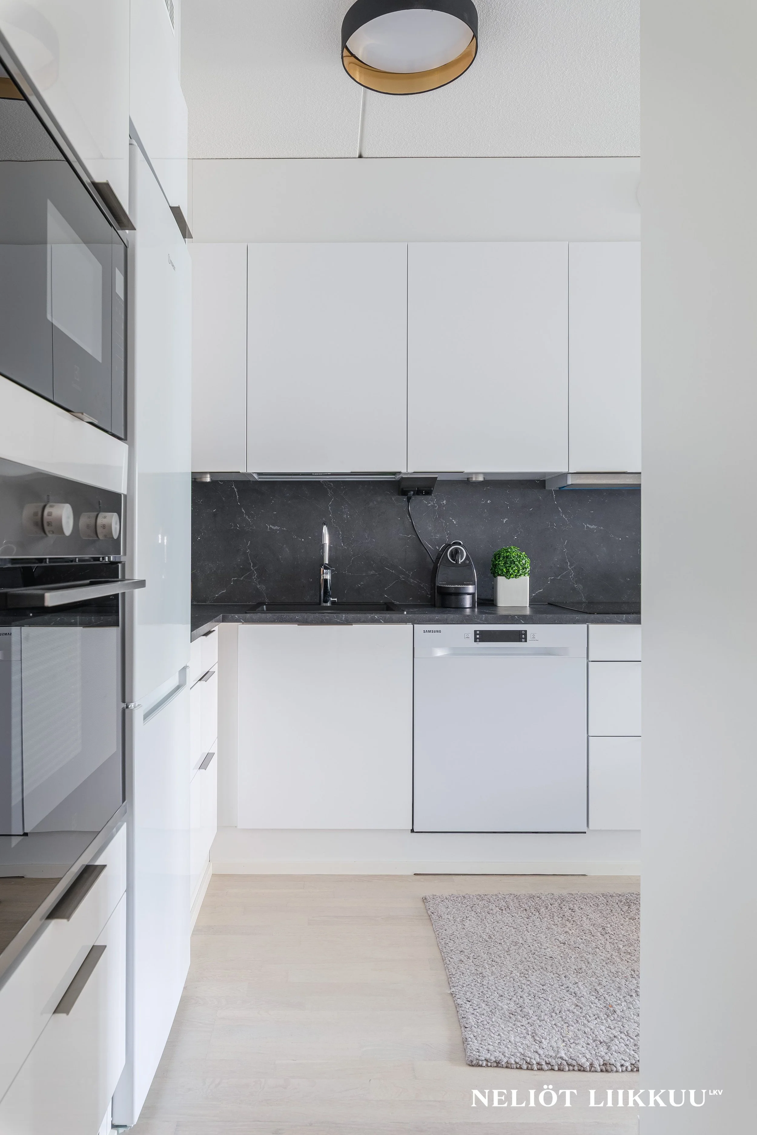 Moderni keittiö, jossa valkoiset kaapistot, musta kivitaso, liesi, astiakaappi ja pienkoneita. Tila on siisti ja minimalistinen.
