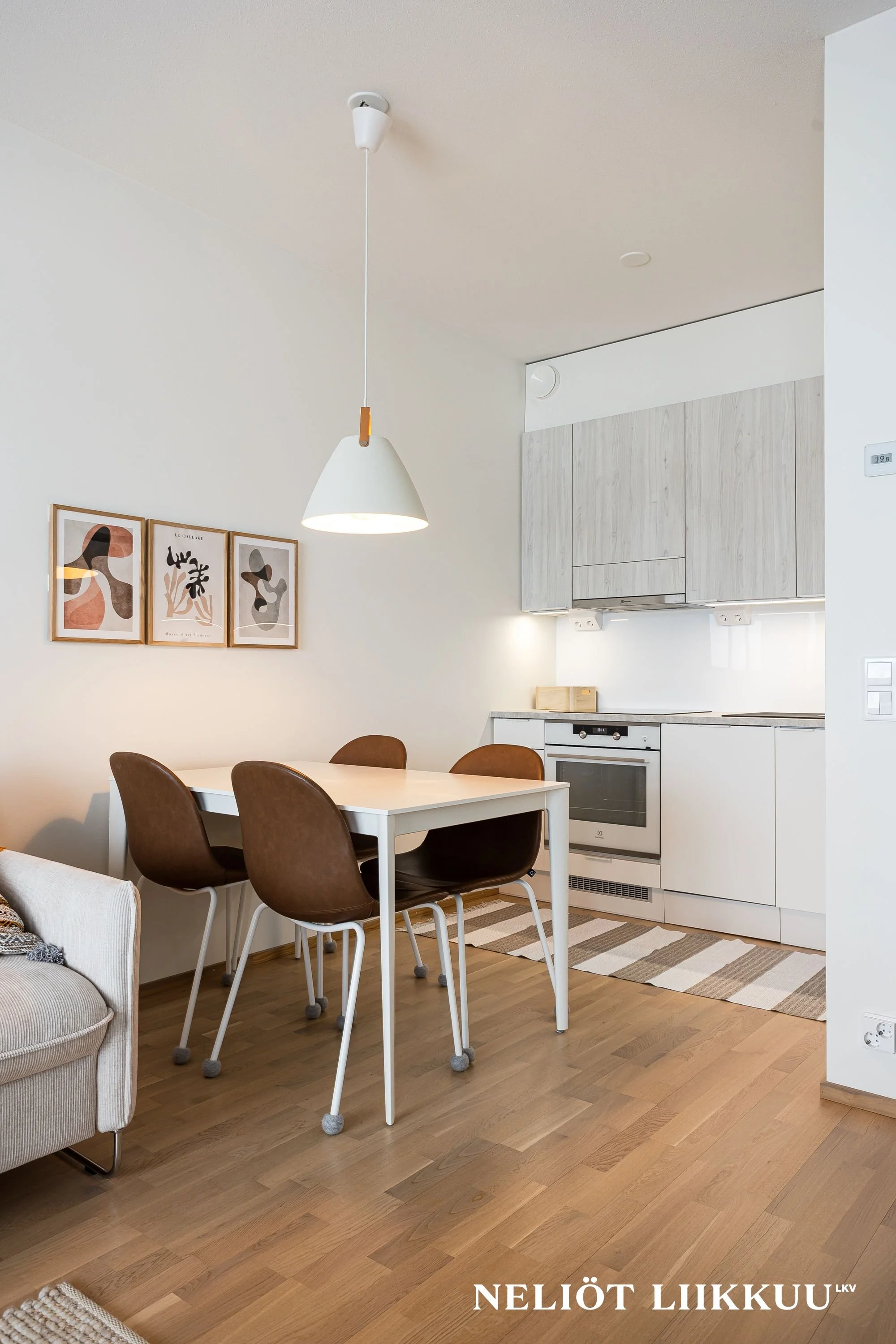 A modern, minimalist kitchen with a white table, brown chairs on wheels, and light wood flooring. There are three abstract art prints on the wall, a hanging pendant light, and light wood cabinets. The scene is bright and tidy.