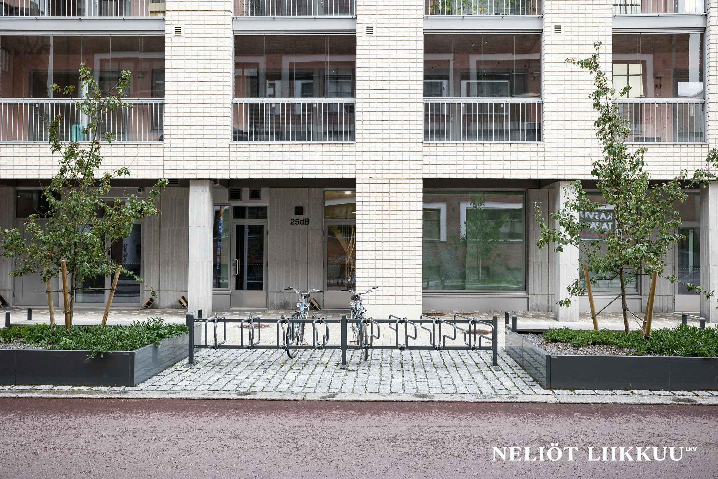 Kuva kerrostalon ulko-ovesta, jossa on pieniä puita ja pyöräparkki edessä, sekä teksti "NELJÖT LIIKKUU" oikeassa Alakulmassa.