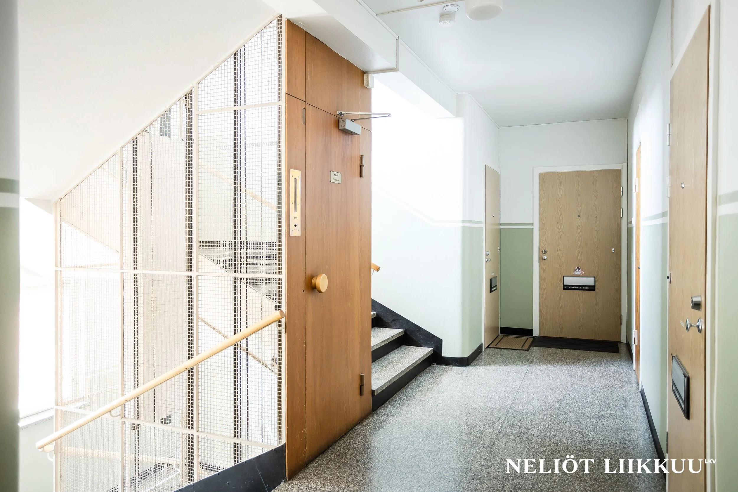 Helsingeissä sijaitseva hissi ja eteinen rakennuksen sisällä, käytävä ja ovi, puiset seinät ja jalka- ja kädensijat.