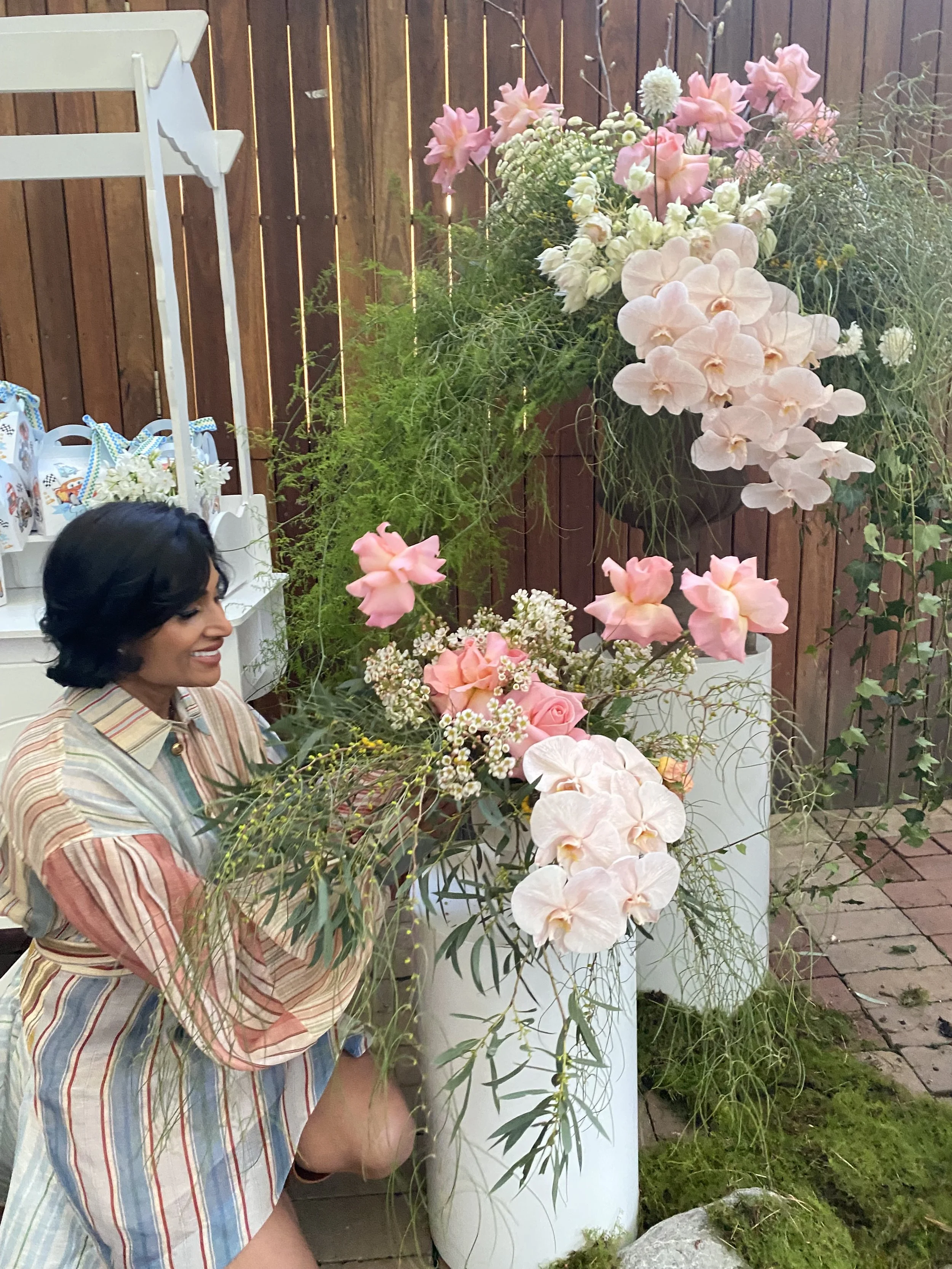 A smiling woman with short dark hair looking at a large colorful flower arrangement in white pots, with pink and white flowers, set against a wooden fence background.