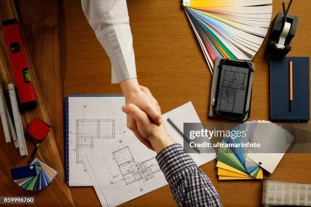 Two people shaking hands over an architectural blueprint on a wooden desk, surrounded by color swatches, a smartphone, notebook, and various stationery items.