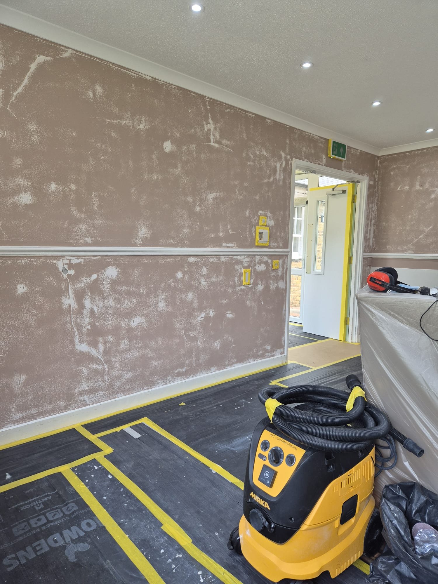 Interior room undergoing renovation with a partially painted brown wall, protected flooring, and yellow painter's tape. A vacuum and equipment are on the floor, and there is an open door leading outside with an emergency exit sign above it.