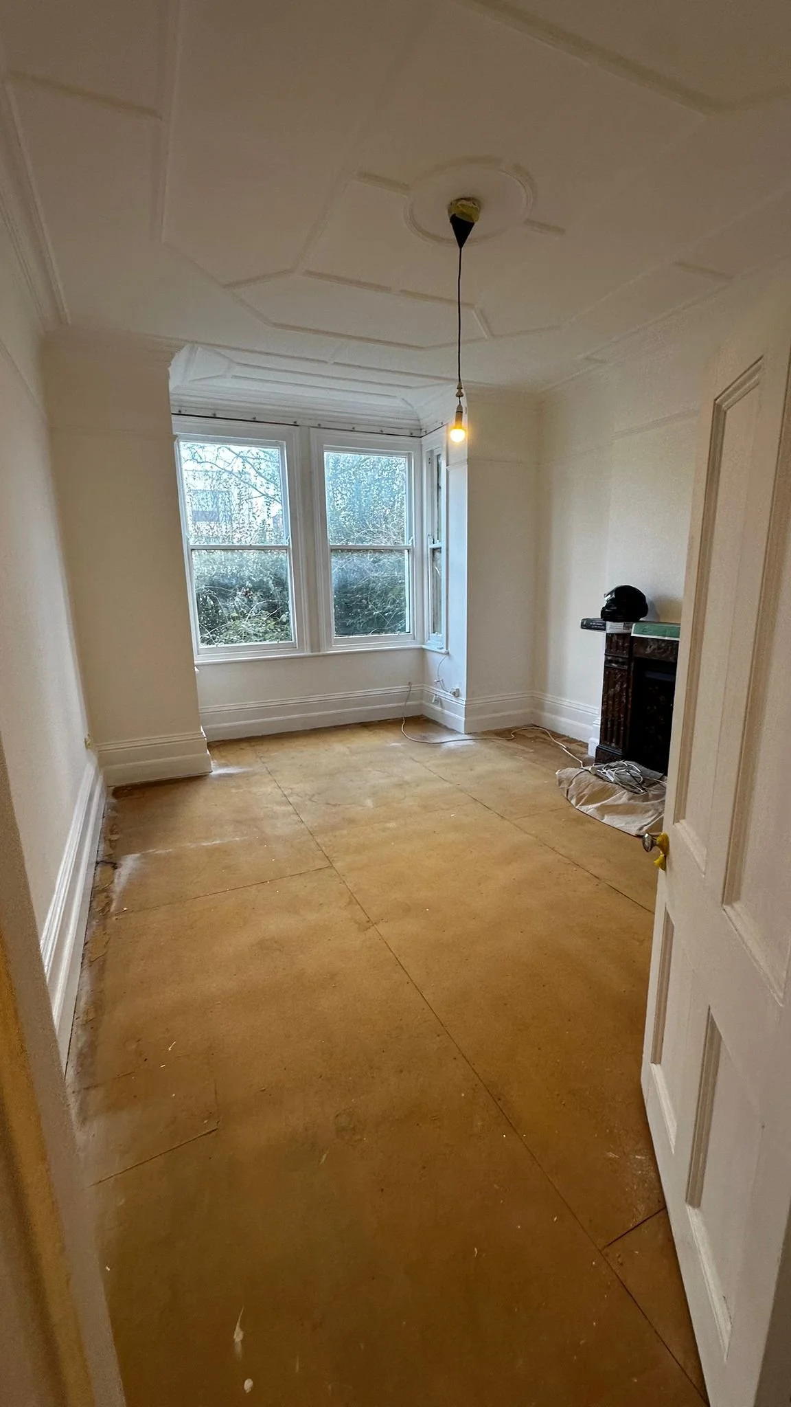 Empty room with a window, high ceiling with decorative molding, unfinished floor, ceiling light fixture hanging, and some items on a small fireplace or shelf.