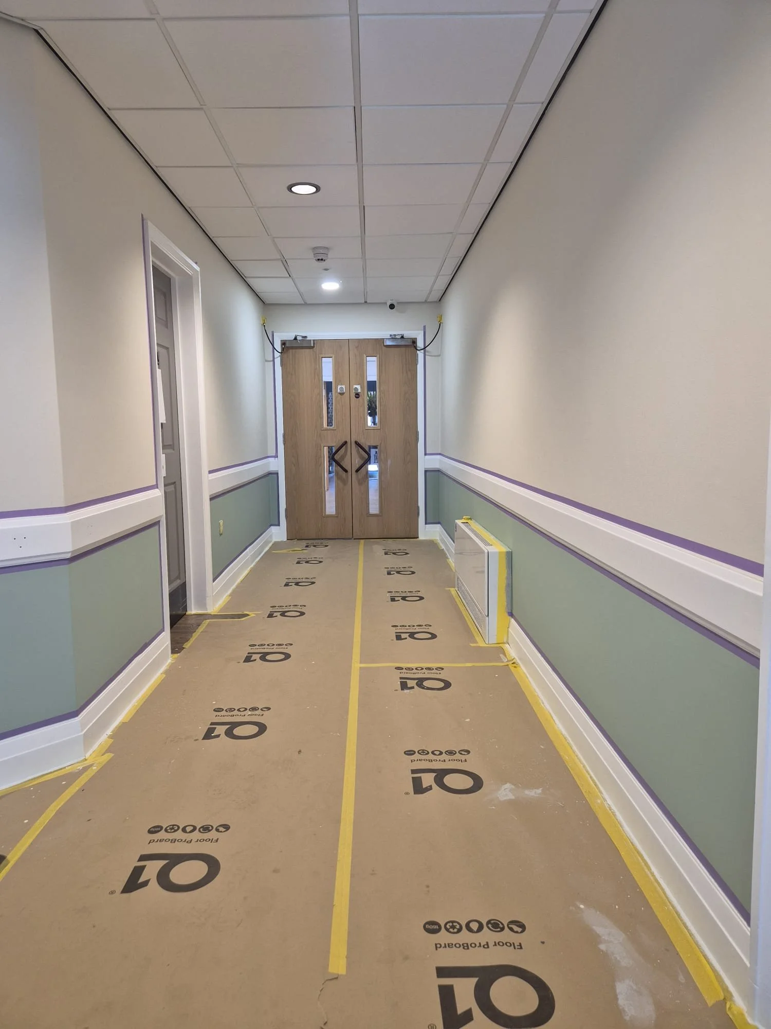 A hallway under construction with new walls painted in light blue and cream, a wooden double door at the end, and yellow tape on the floor marking the edges of the underlayment. The ceiling has white tiles with a few lights and smoke detector.