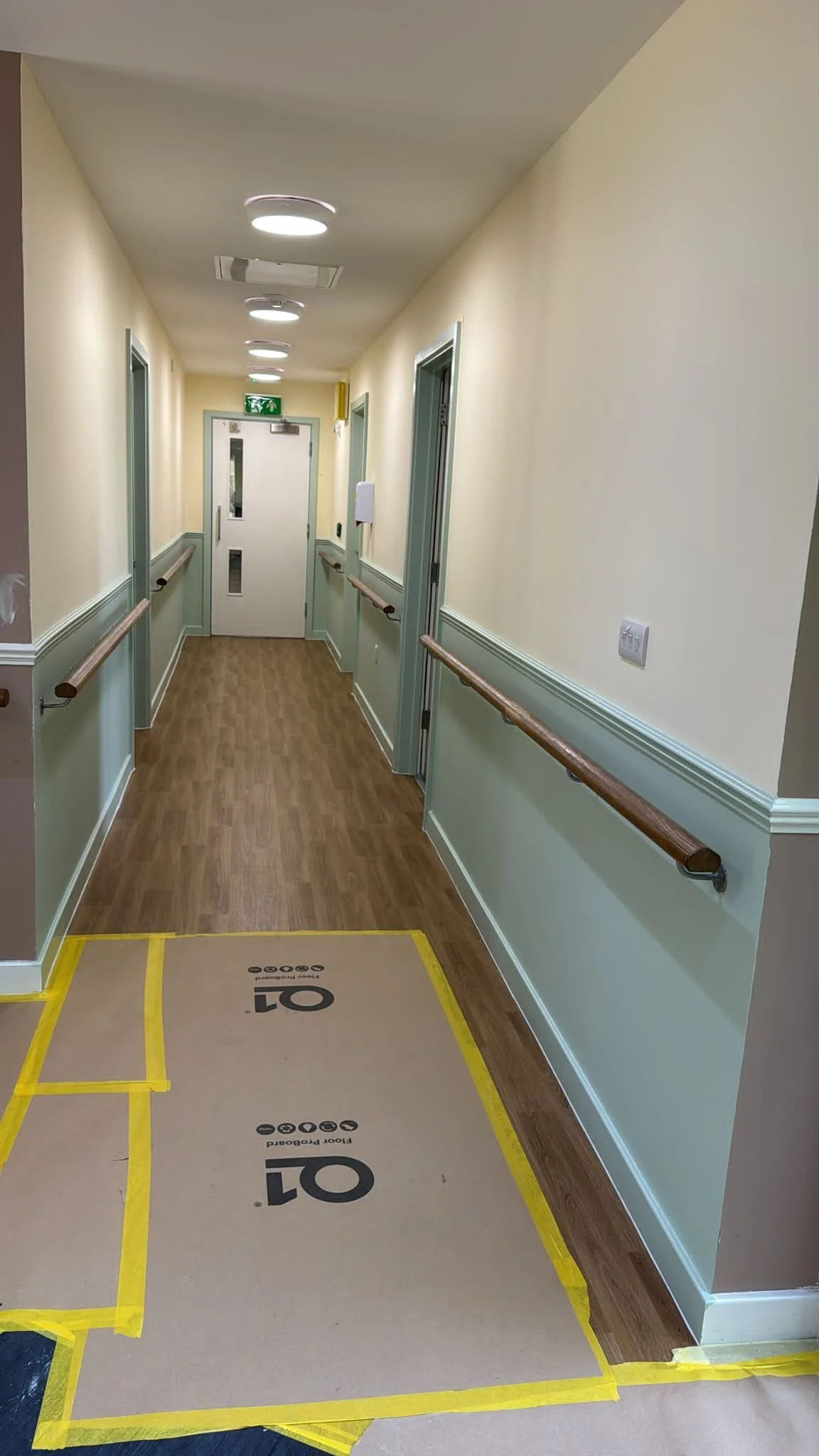 Hospital corridor with yellow and beige walls, wood flooring, emergency exit sign, and a floor protection covering marked with '01' in black.