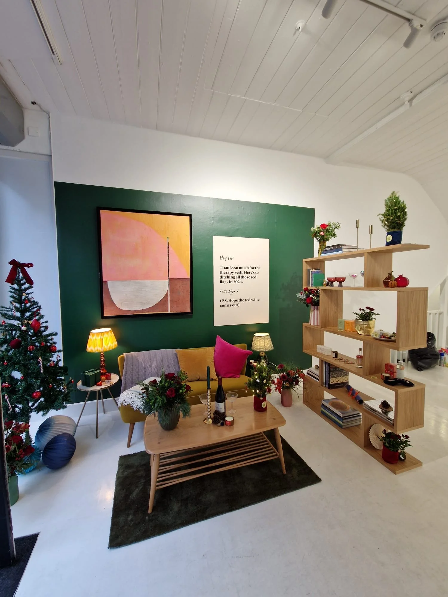 A cozy living room decorated for Christmas with a Christmas tree, assorted ornaments, flowers, candles, books, and framed art on green-painted wall.