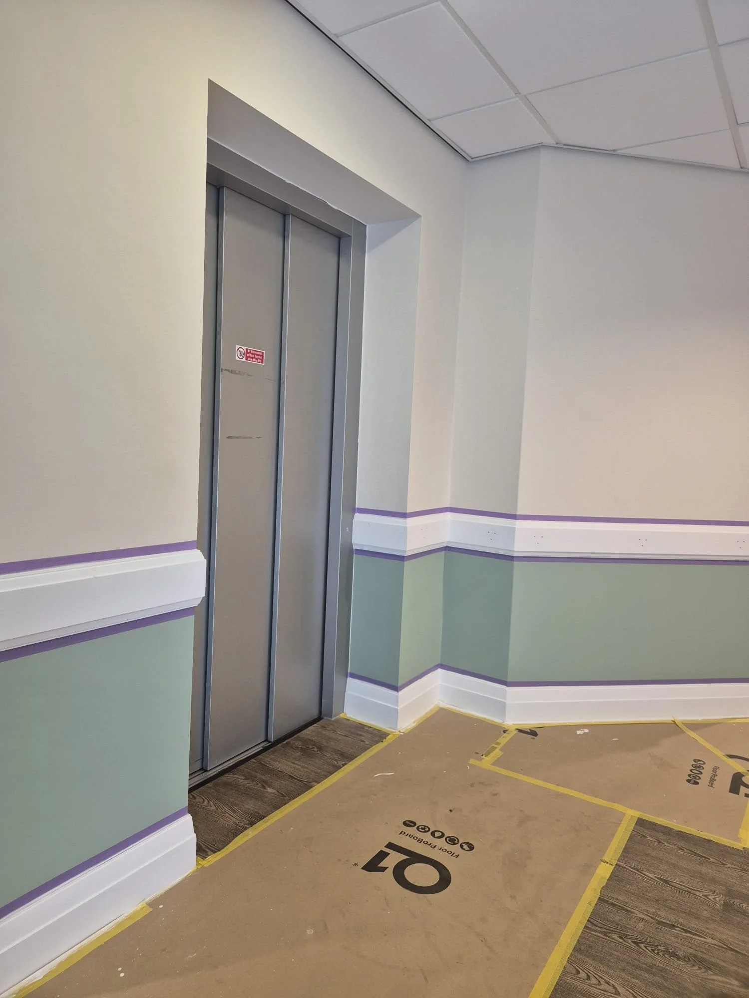 Interior of a building under construction with elevator door, painted walls with purple and green stripes, and protective floor covering.
