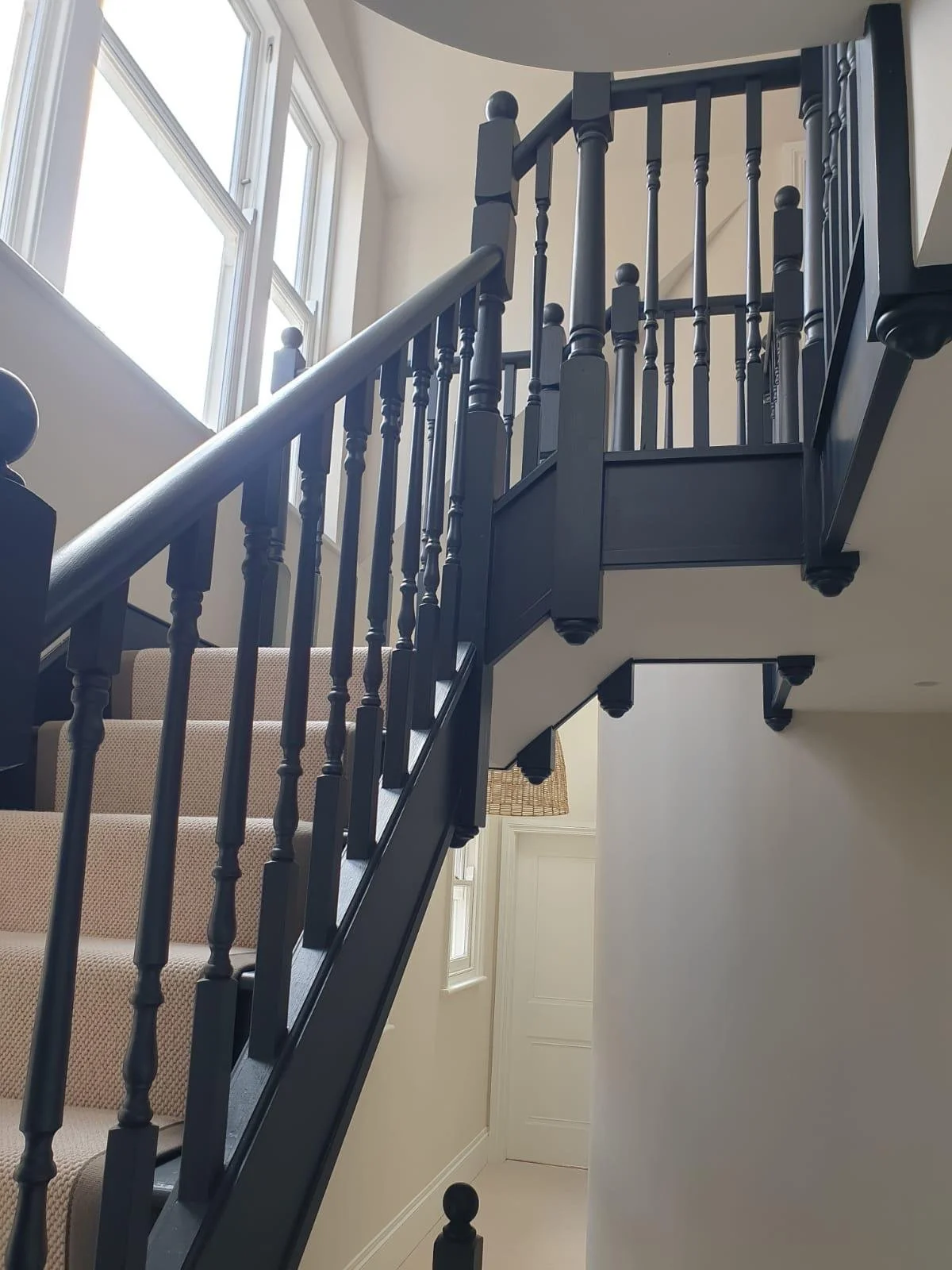 Interior view of a staircase with dark wood railings and beige carpeted steps in a well-lit home, with large windows and white walls.