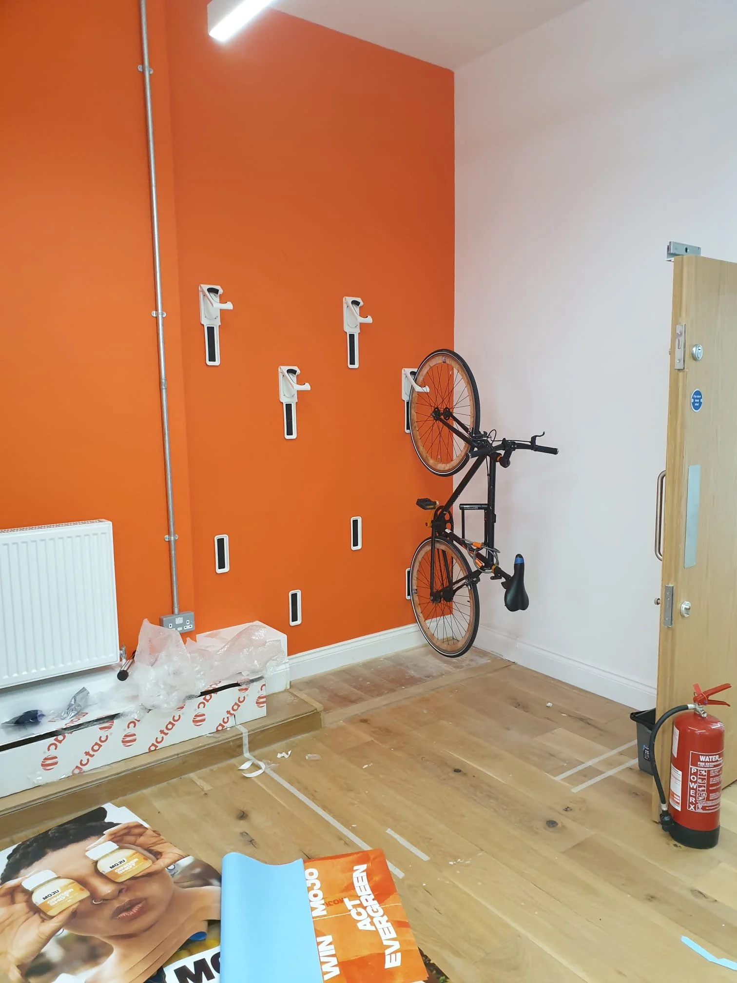 An indoor room under renovation with an orange wall and a white wall, bike against the orange wall, fire extinguisher, and miscellaneous items on the wooden floor including a large poster or brochure with a woman holding two yellow jars.