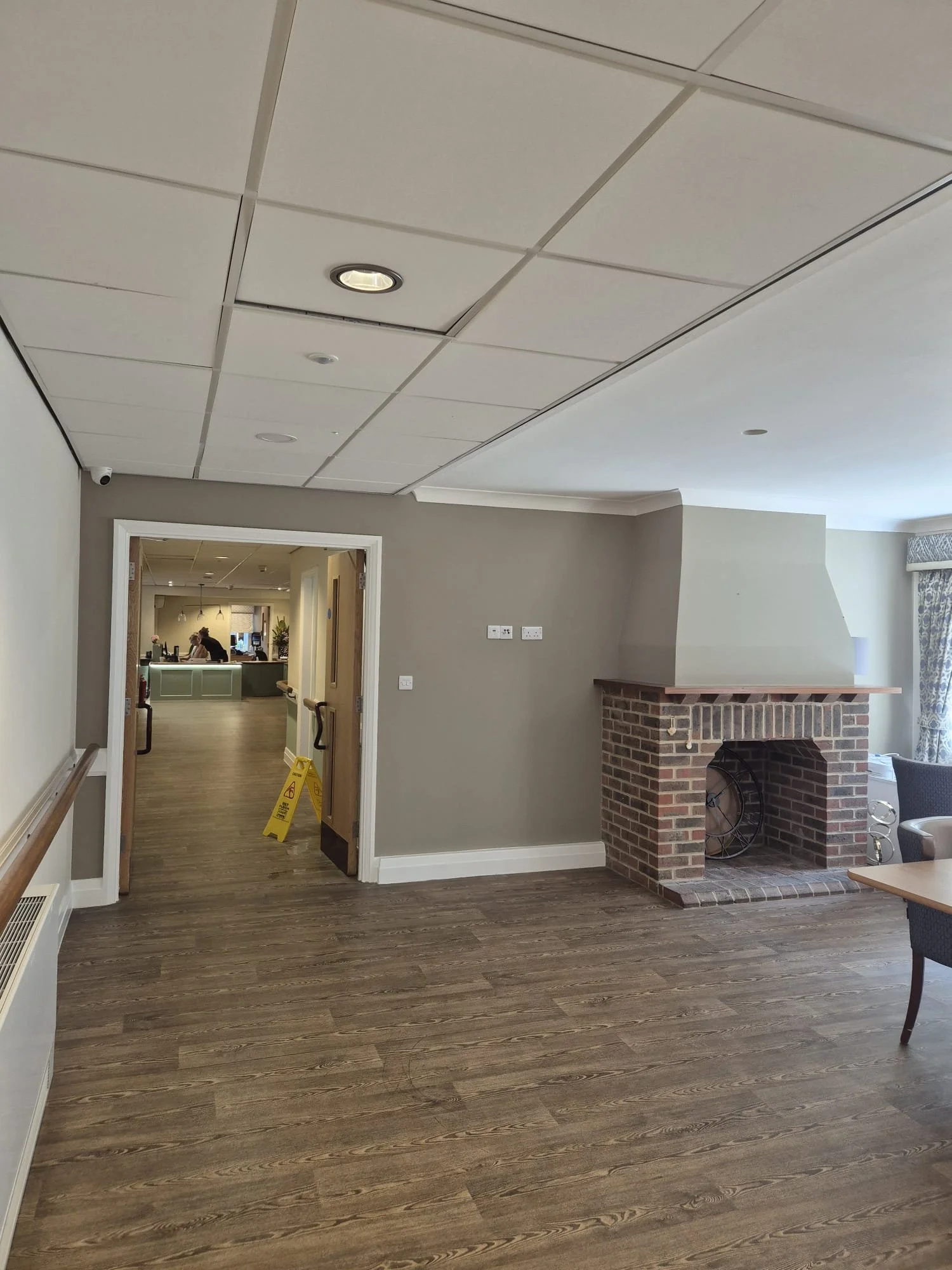 A room with wood flooring, beige walls, a brick fireplace, and a doorway leading to another room. There is a chair partially visible on the right, a yellow caution sign on the floor, and ceiling lights. The other room has people working and a desk vi
