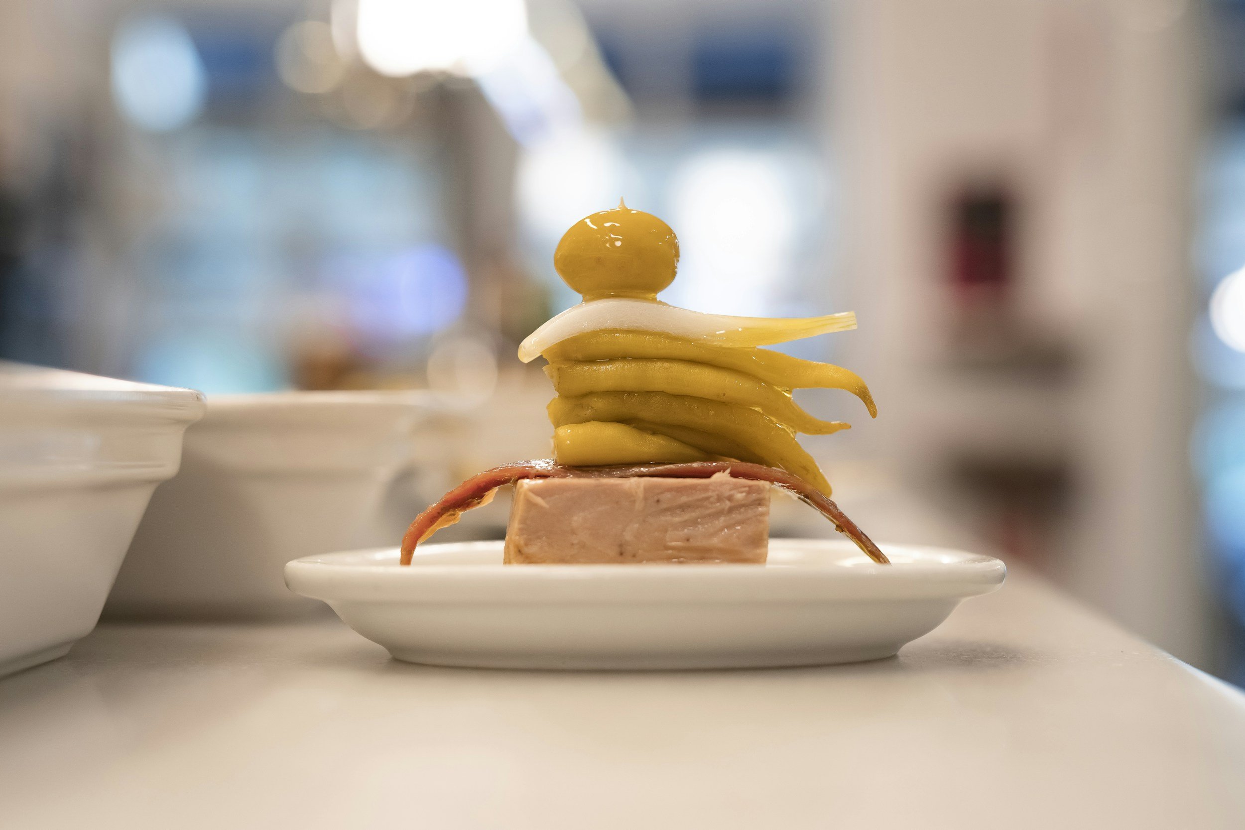 Stacked appetizers on small white plates, including slices of cured meat, a slice of cheese, artichoke hearts, and a small piece of mozzarella with a green olive on top, set against a blurred background.
