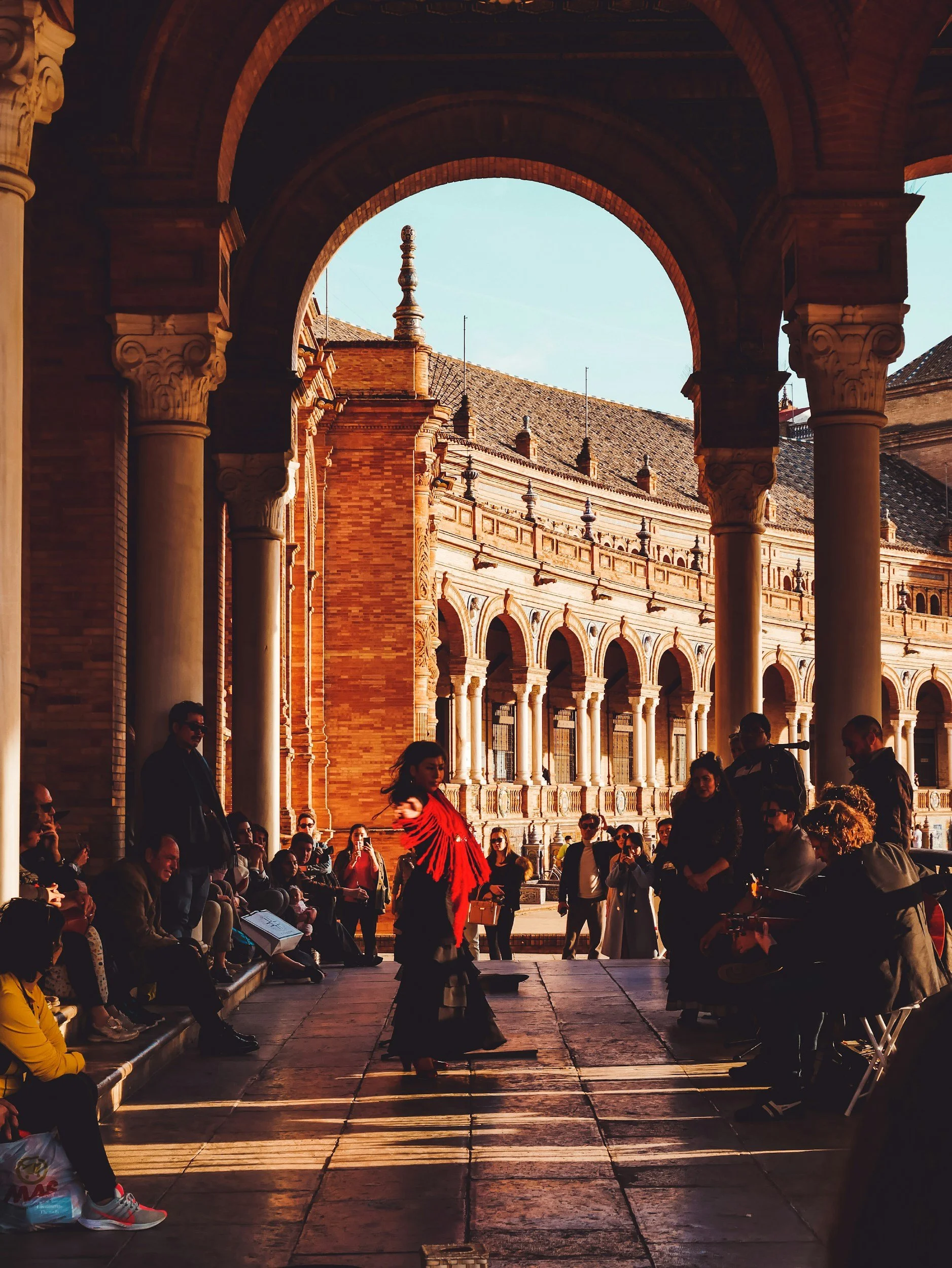 A Spanish flamenco dancer performing outdoors under an arched corridor with striped flooring, with an audience seated and standing, and a building with ornate architecture in the background.