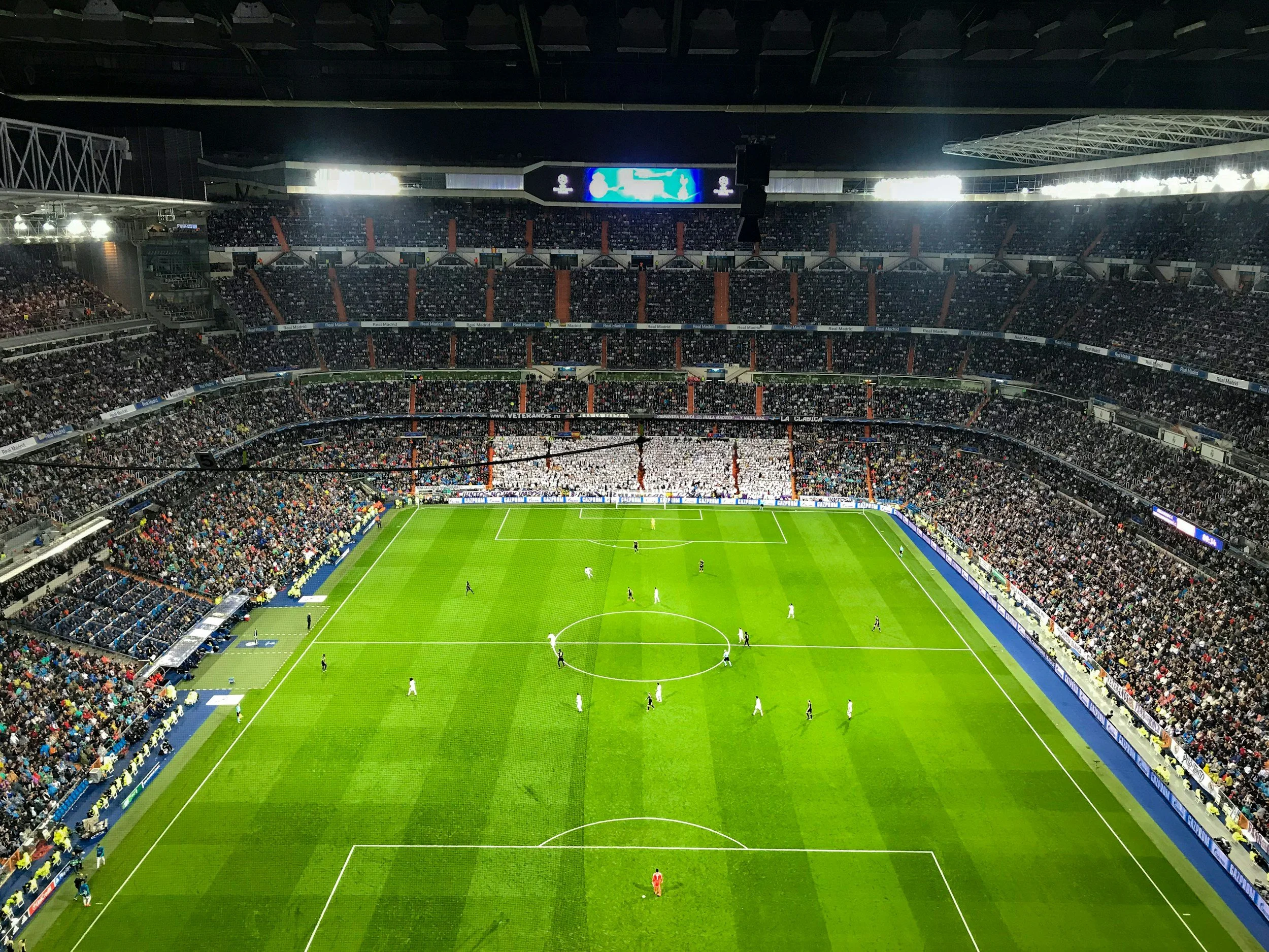A packed Spanish soccer stadium with a green pitch and players visible, viewed from a high angle during a night match.