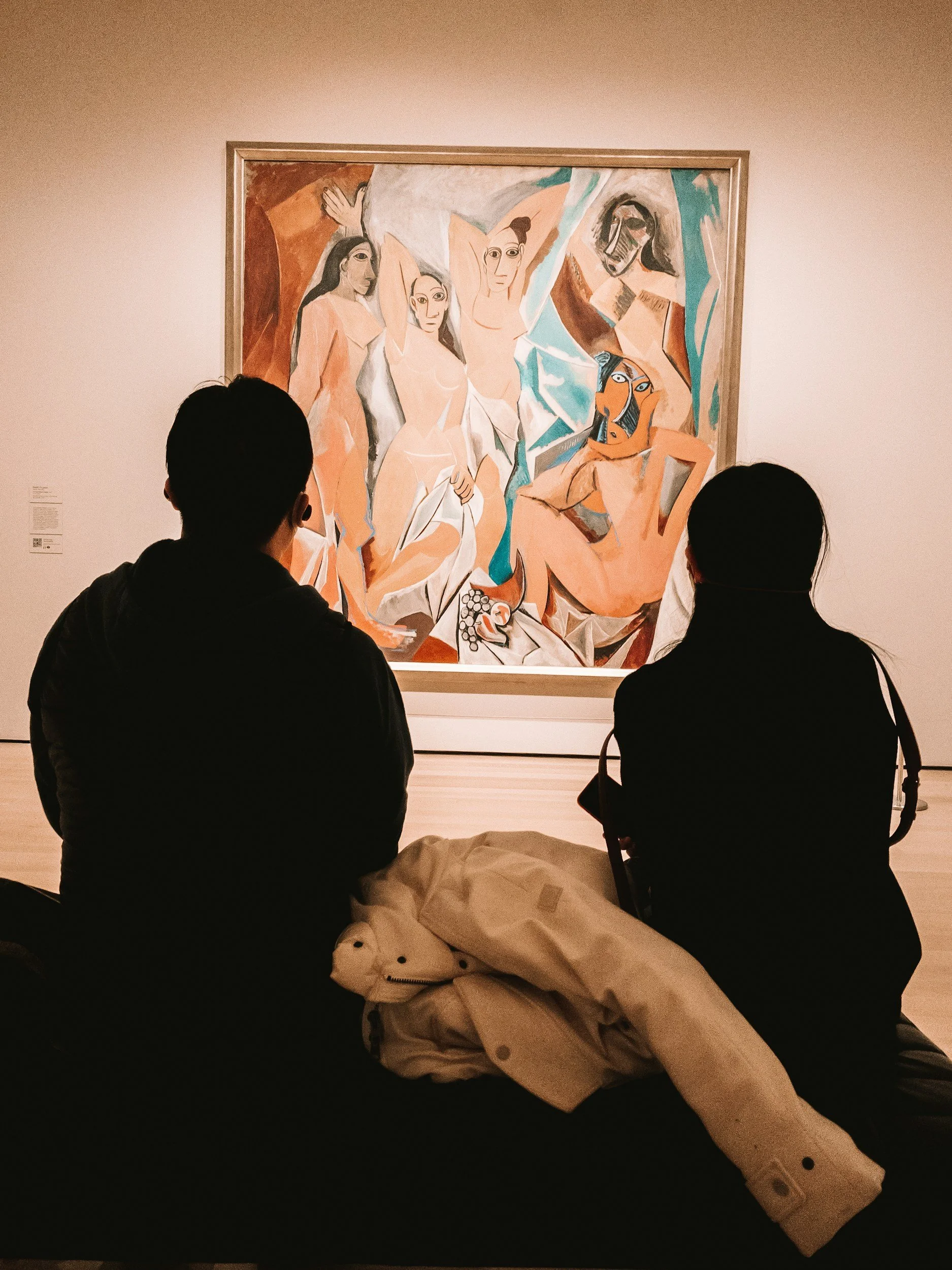Two people seated in front of a painting with abstracted human figures in an art gallery.