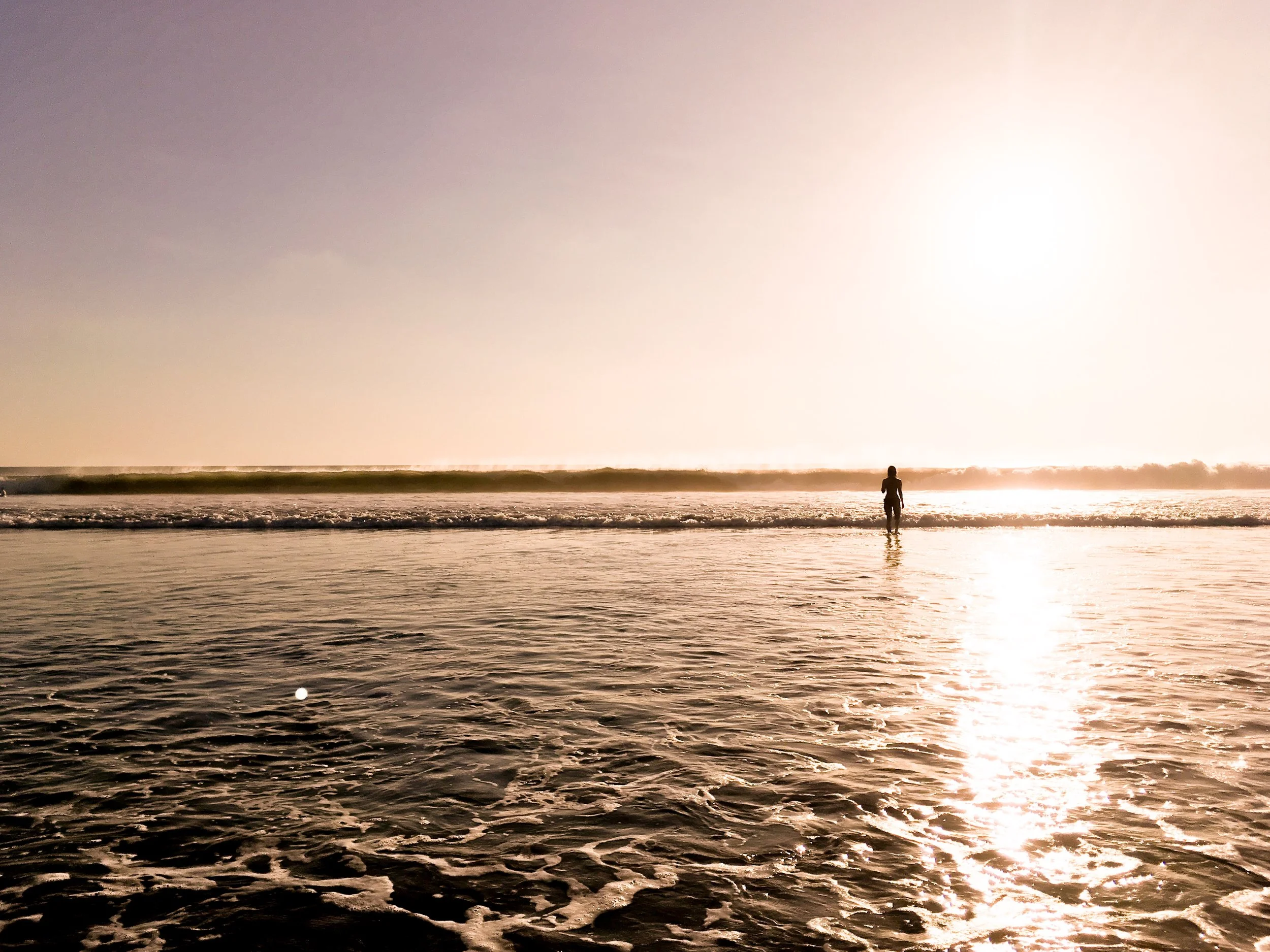 Ein einzelner Mensch steht am Strand im Wasser während des Sonnenuntergangs, der Himmel ist hell und die Sonne spiegelt sich im Wasser wider.