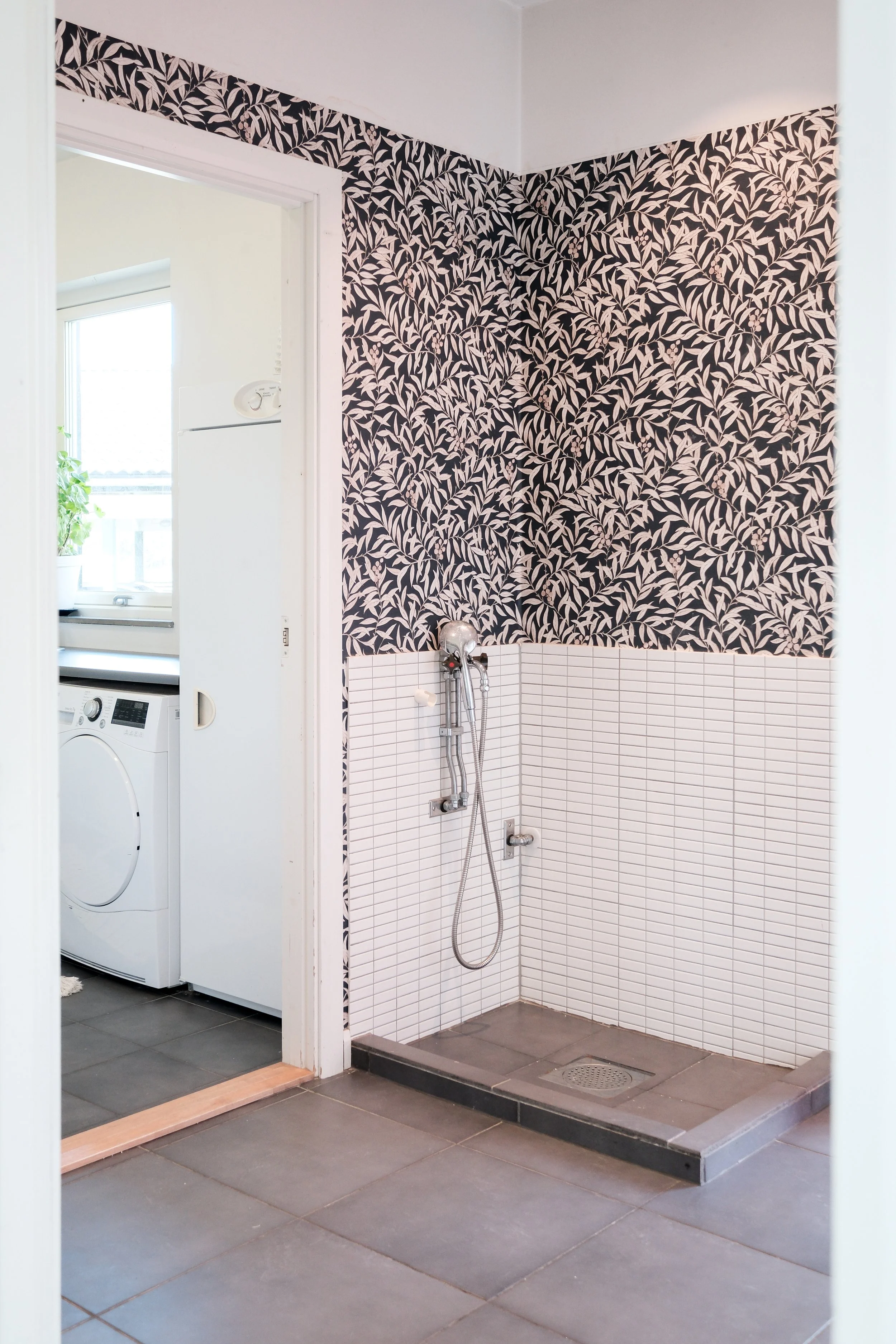A walk-in shower area with white tiled walls, a handheld showerhead, and a dark gray tiled floor. To the left, there is a small step with tile transition to a room with dark gray floor tiles where a laundry area with a washing machine and a dryer is 