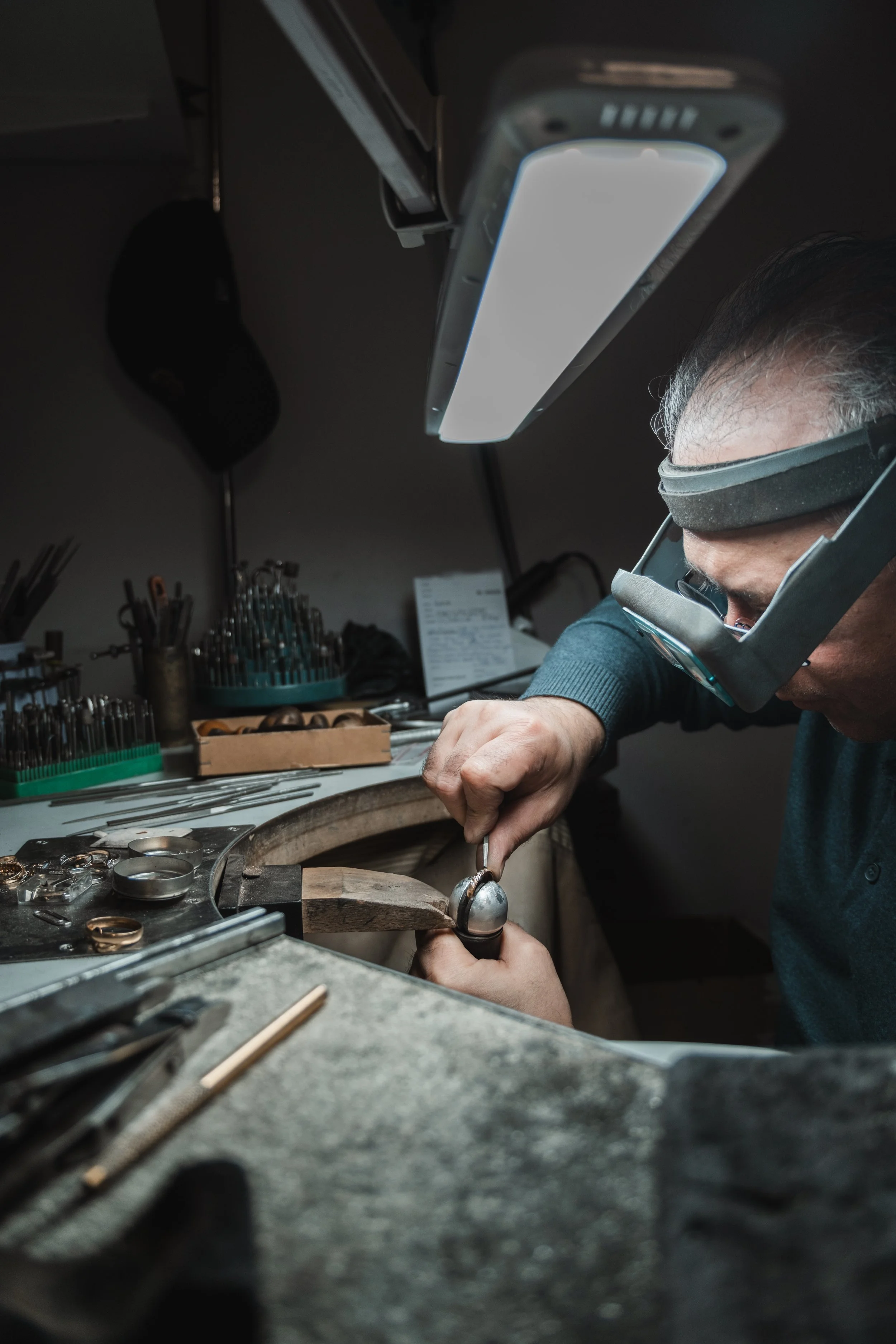 A person wearing magnifying glasses working on jewelry, possibly a ring, with various tools and jewelry pieces on a workbench underground workshop.
