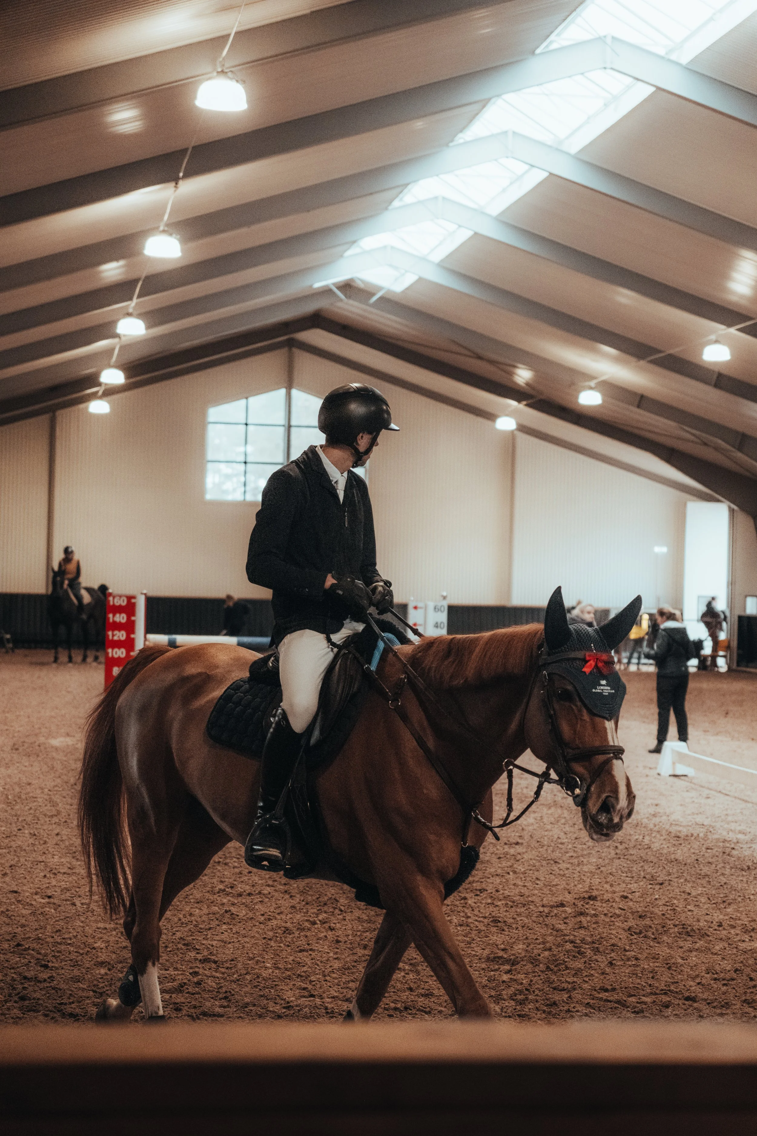 A person wearing a helmet and riding a horse inside an indoor arena with a high vaulted ceiling and skylights. Other people are present in the background.