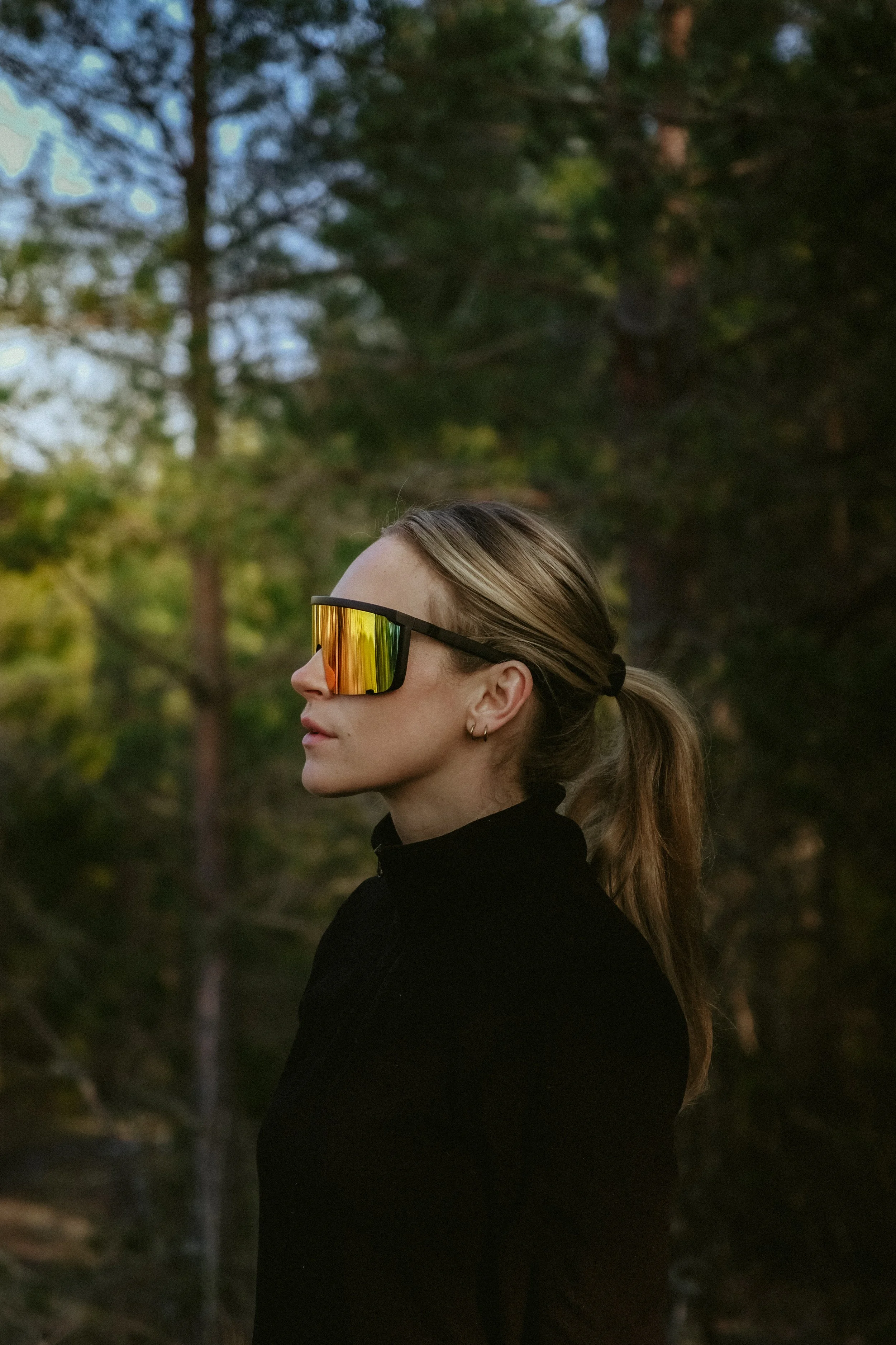 Side profile of a young woman with light brown hair tied in a ponytail, wearing reflective sunglasses and a black top, standing outdoors with a blurred background of trees and sky.