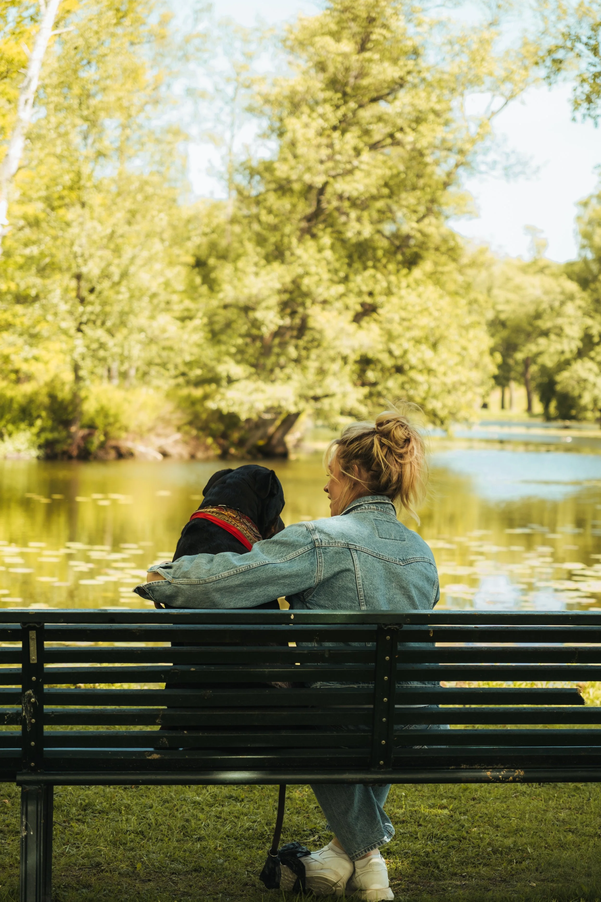 A young woman with blonde hair sitting on a black park bench with a black dog, near a body of water in a park with green trees and blue sky in the background.