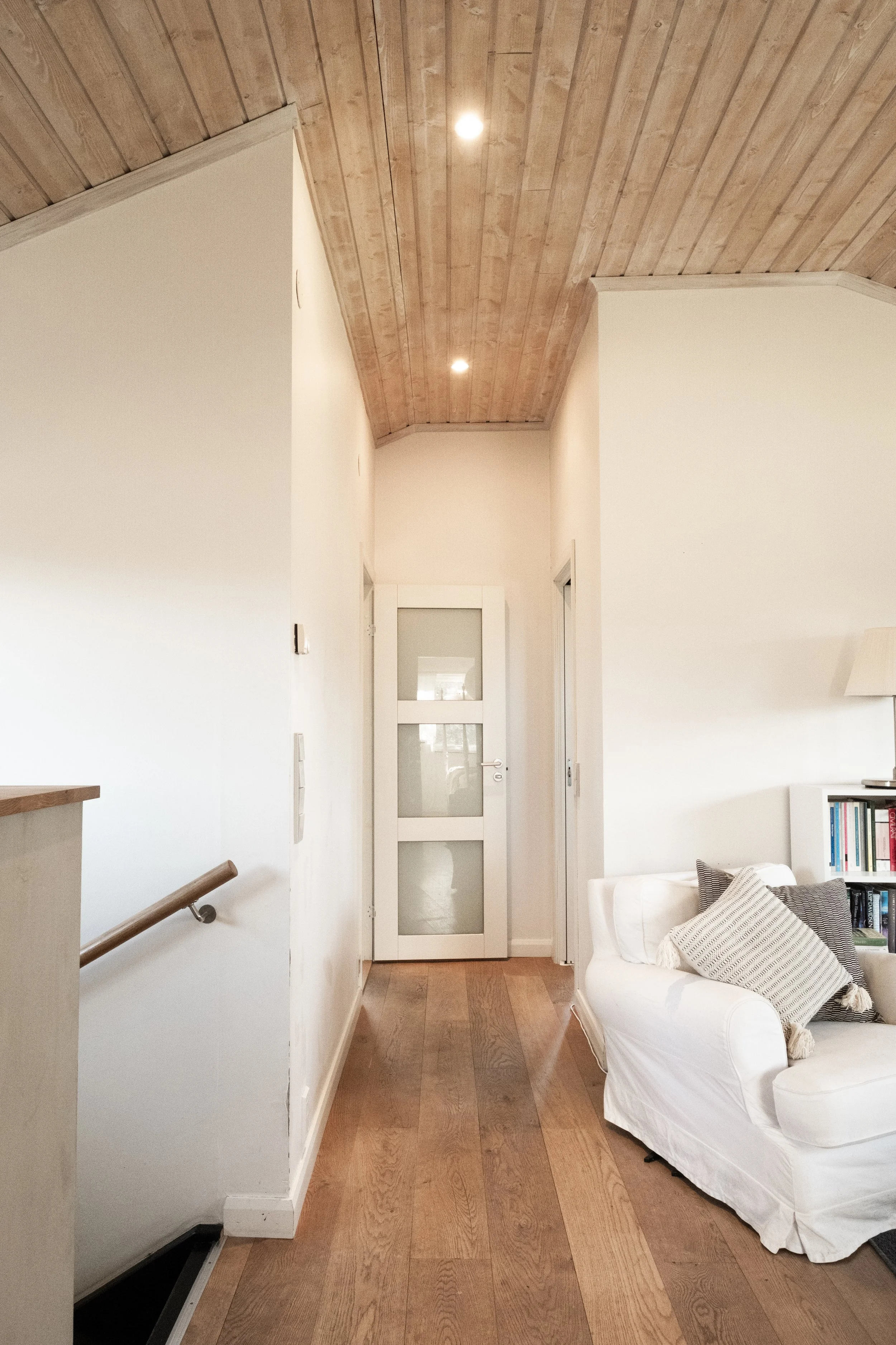 Interior view of a hallway with wooden floors and a wood-paneled ceiling, white walls, a white door at the end, a bookshelf with books on the right, and a white couch with cushions on the right side.