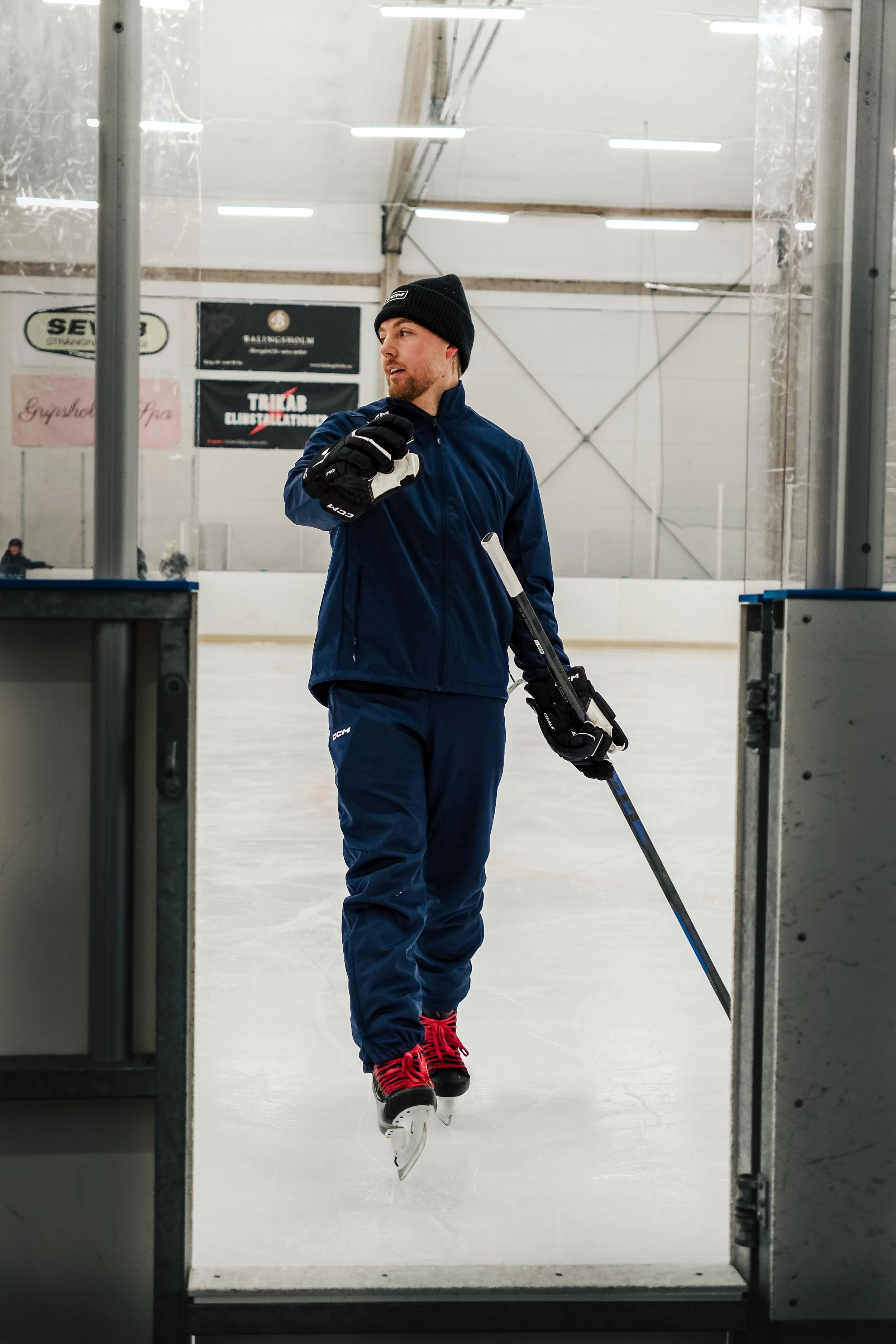 A man in a navy blue winter jacket, black beanie, and red ice skates is standing on the ice, holding a hockey stick, inside an indoor ice rink.