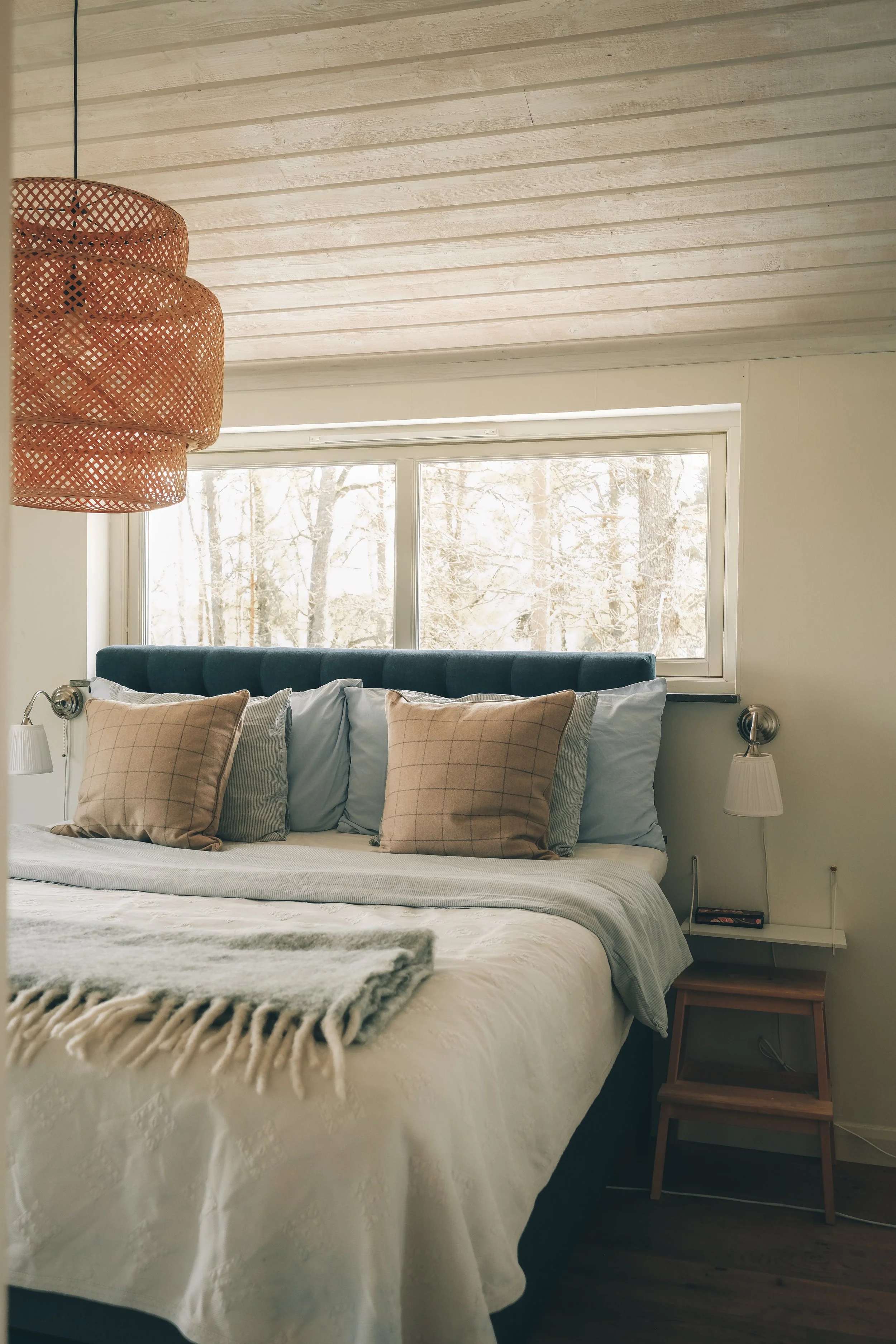 A cozy bedroom with a bed covered in white embroidered bedspread, topped with beige and blue pillows, next to a window showing winter trees, with a wooden side table, a white lampshade, and a woven pendant light.