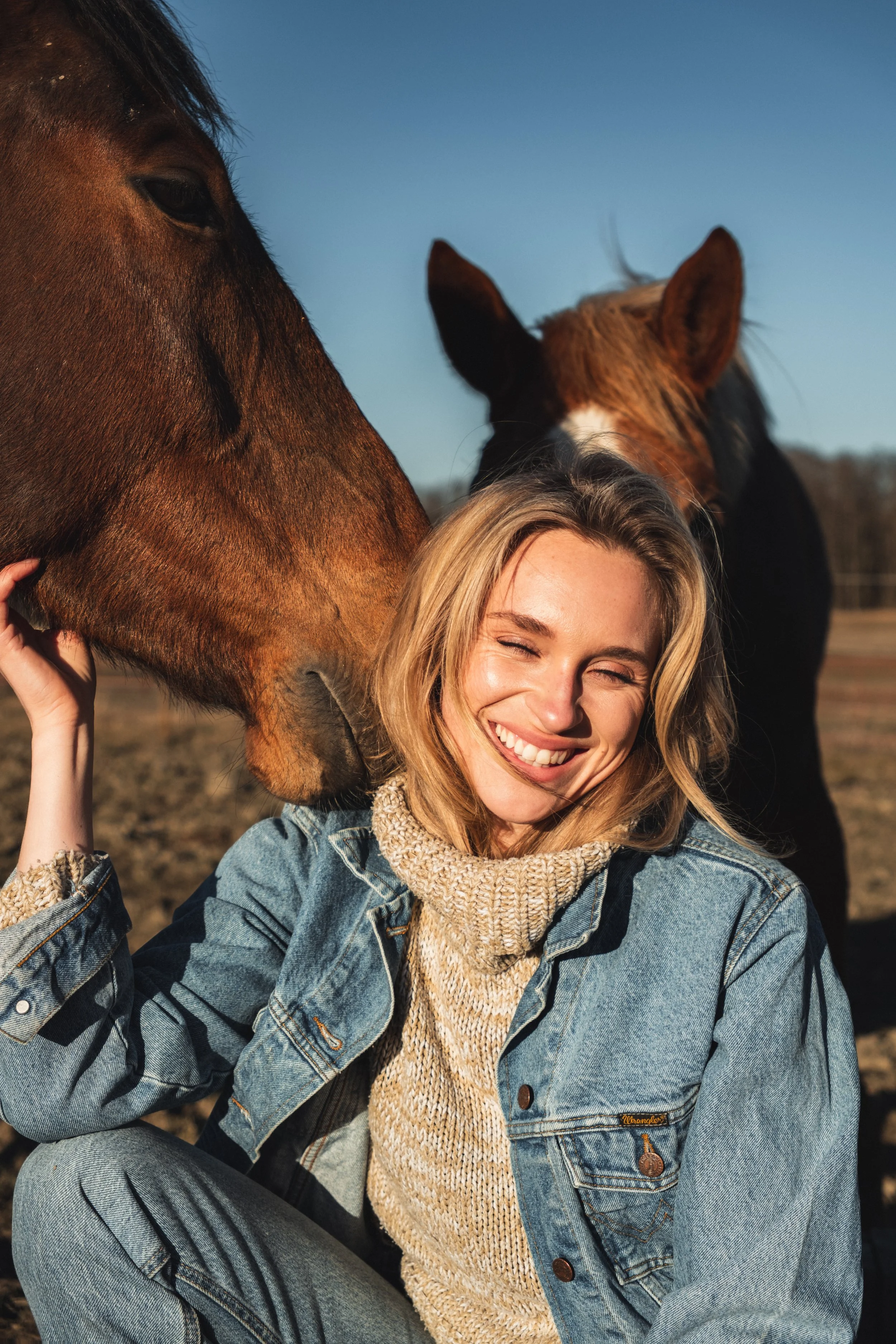 A young woman with blonde hair smiling and squinting, sitting outdoors in sunlight, wearing a denim jacket and a knitted beige turtleneck, with two horses in the background, one brown and one chestnut, under a clear blue sky.
