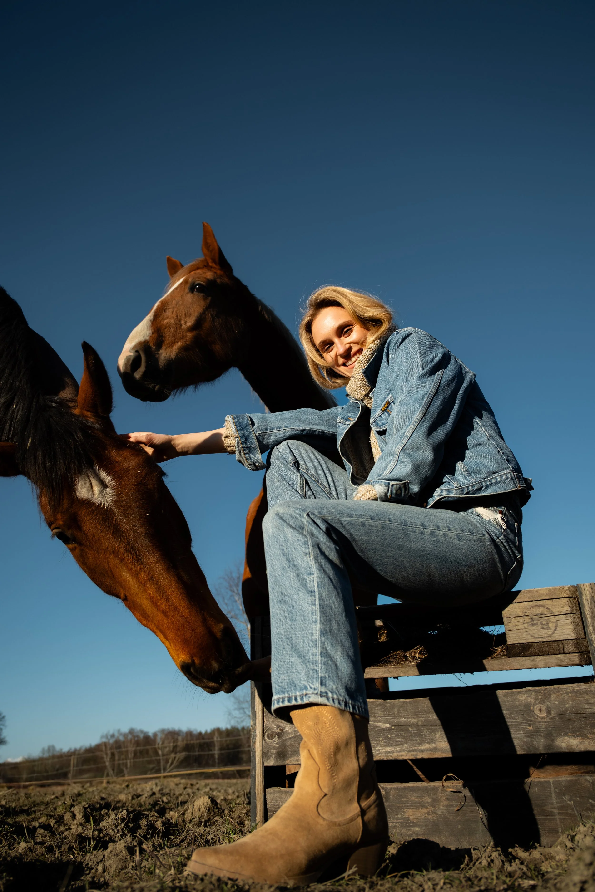 A woman with blonde hair smiling and wearing a denim jacket and jeans, sitting on a wooden fence with two horses, one brown and white, in a field under a clear blue sky.