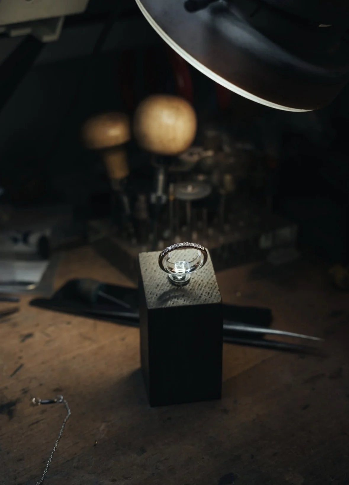A diamond engagement ring resting on a black box in a workshop with tools and machinery in the background.