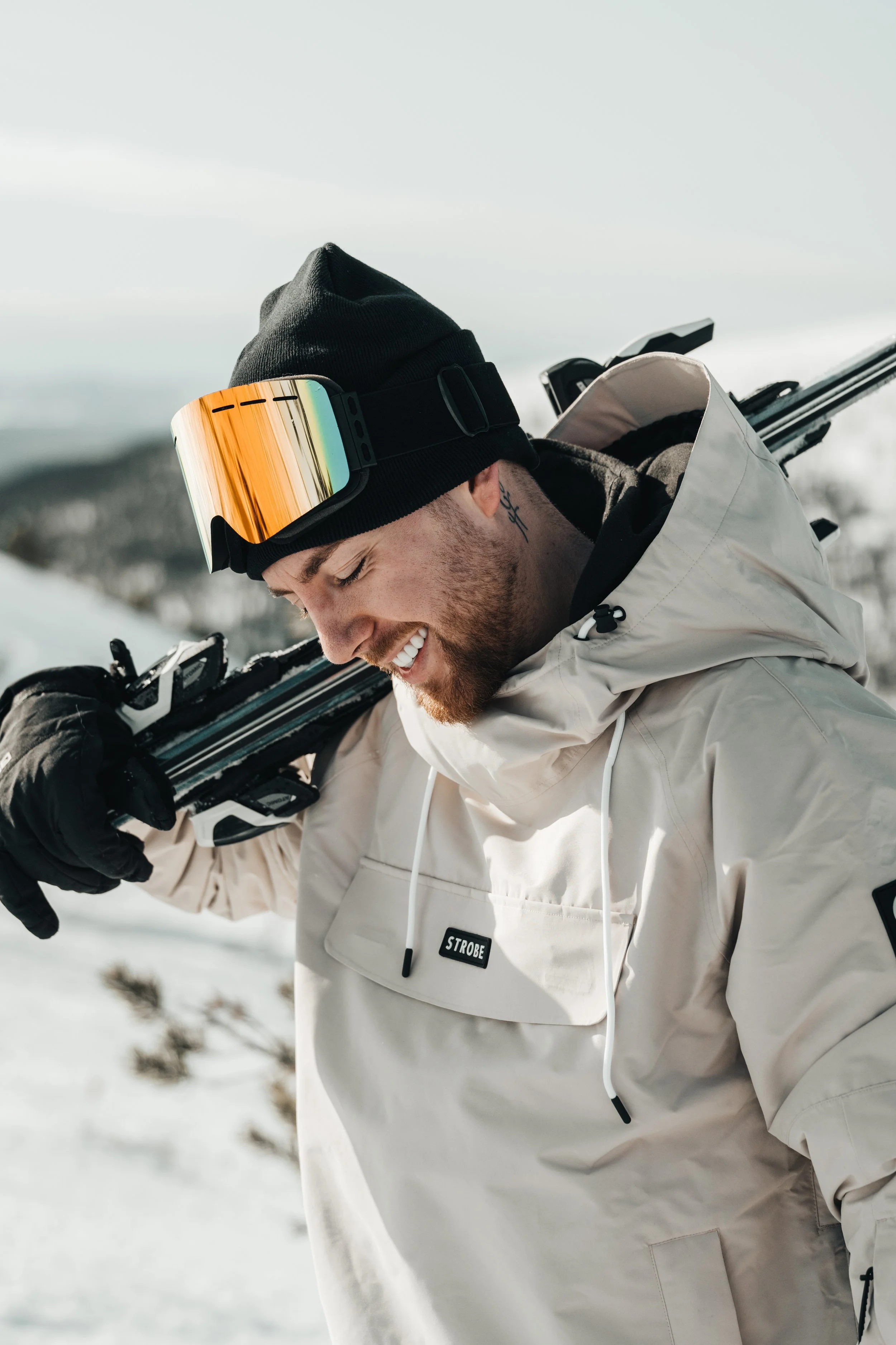Man smiling with closed eyes outdoors in winter, wearing a black beanie, ski goggles, white jacket, and carrying skis on his shoulder.