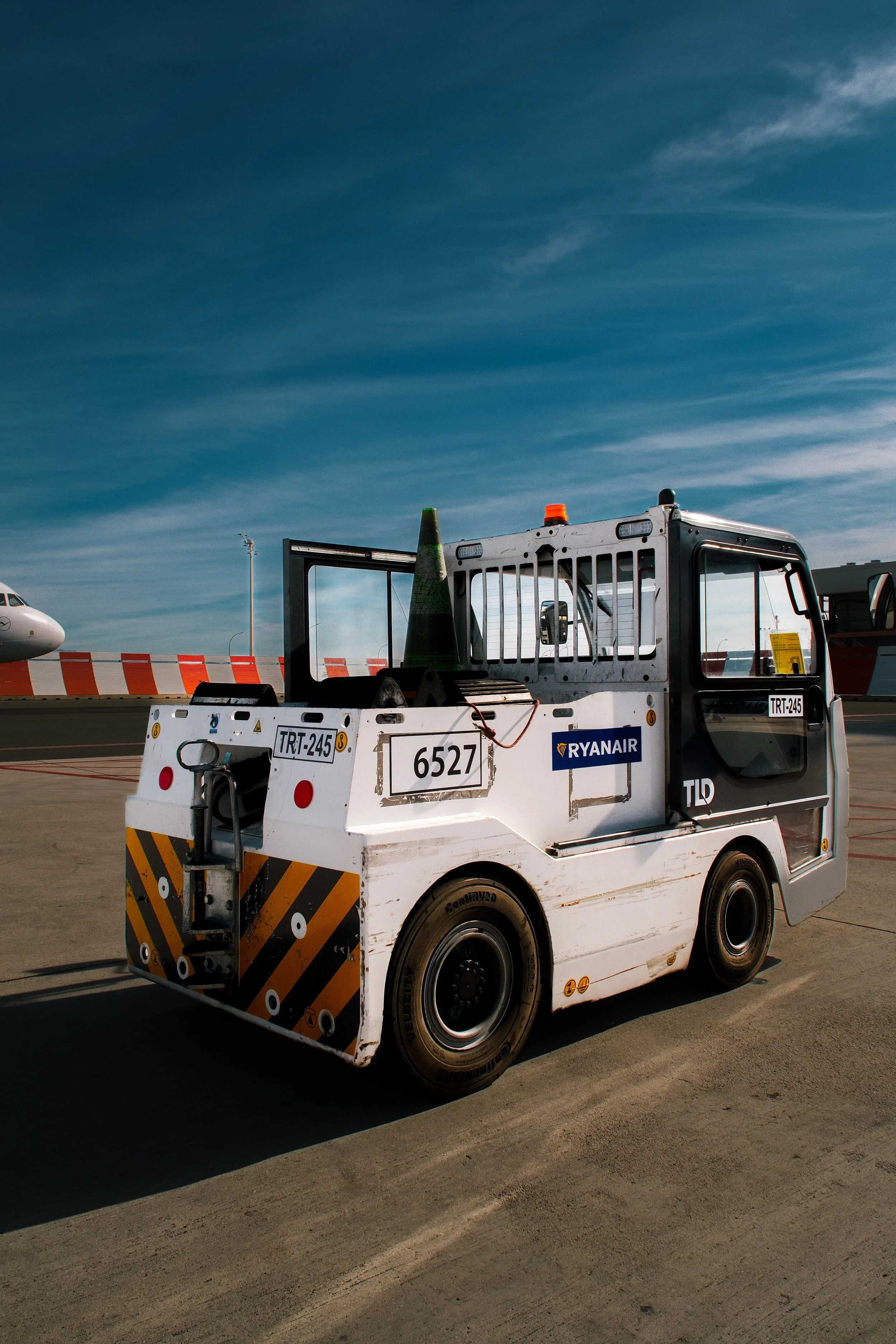 Airport ground crew vehicle with a traffic cone and safety barriers, with airplane in the background on a tarmac under a blue sky with wispy clouds.