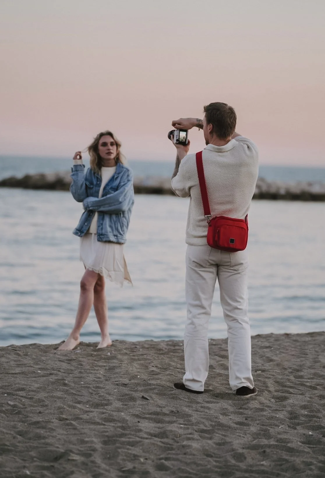A man with a red bag taking a photo of a woman on the beach at sunset.