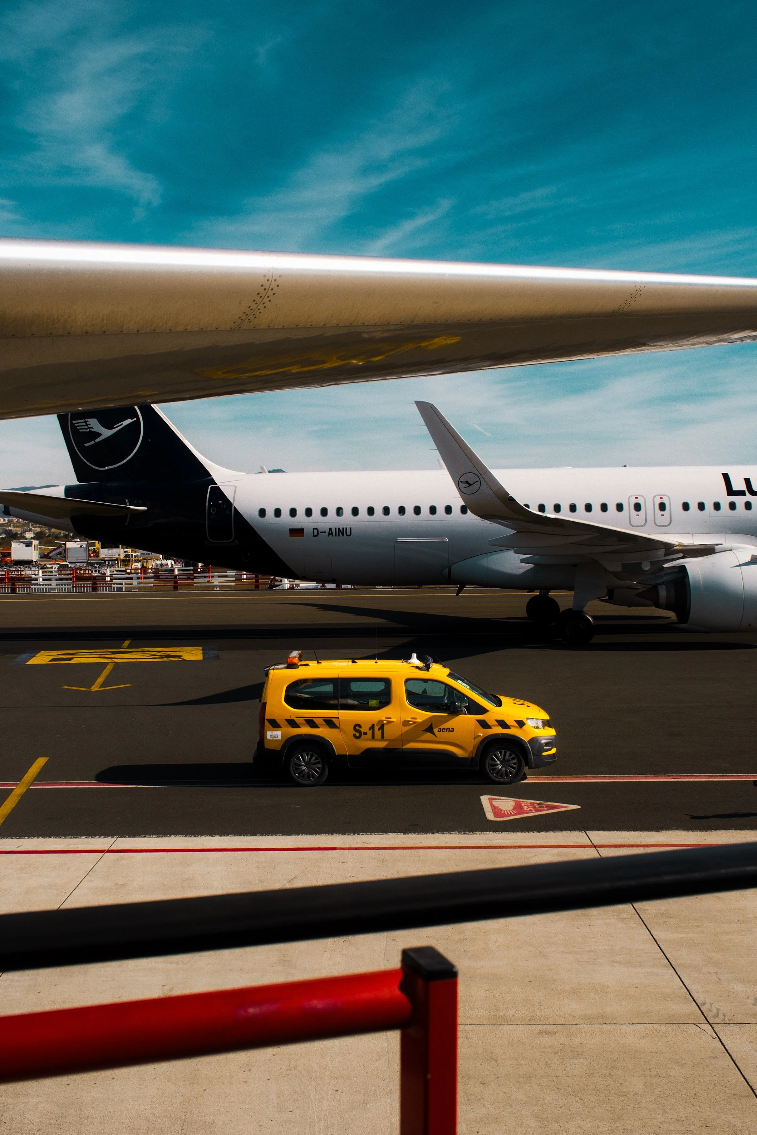 View of an airport tarmac with a Lufthansa airplane, a yellow airport vehicle, and part of an airport terminal. The sky is blue with some clouds.