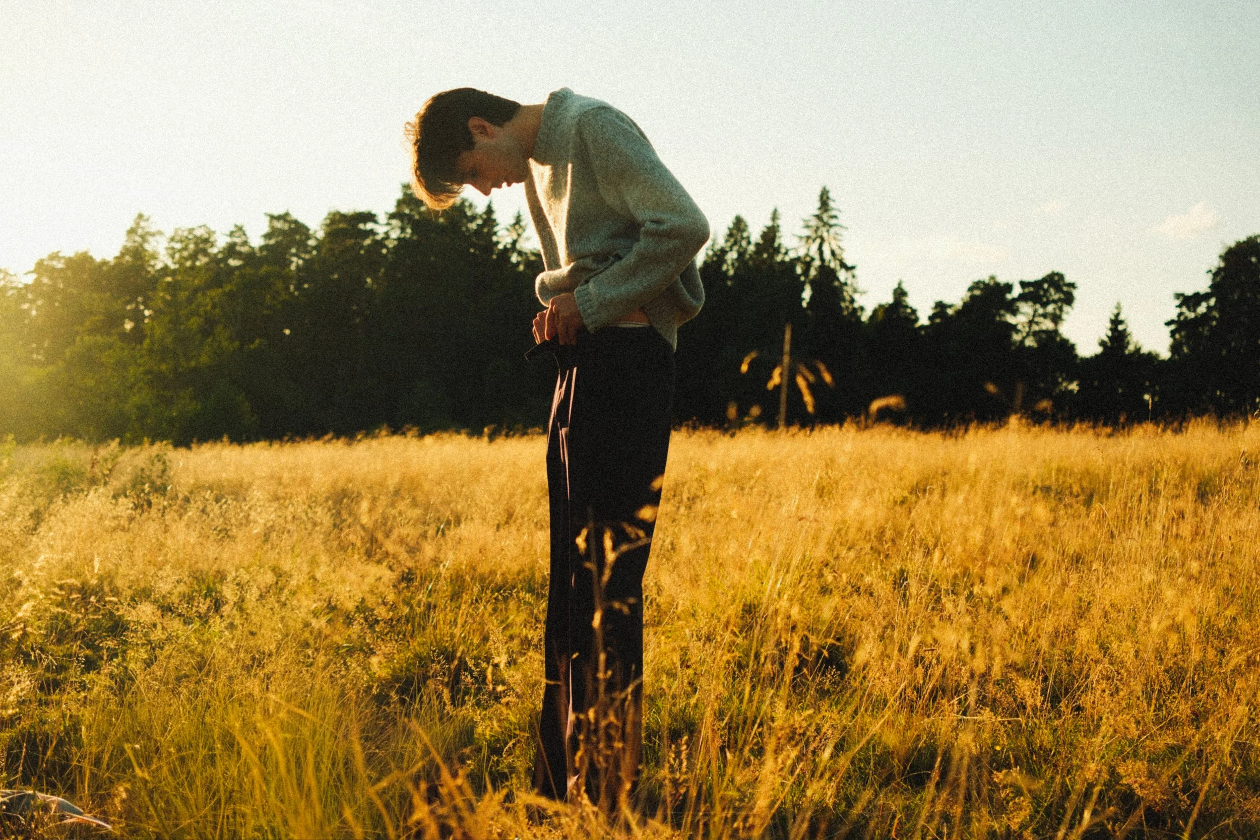 A young man standing in a field of tall grass during sunset, adjusting his pants.