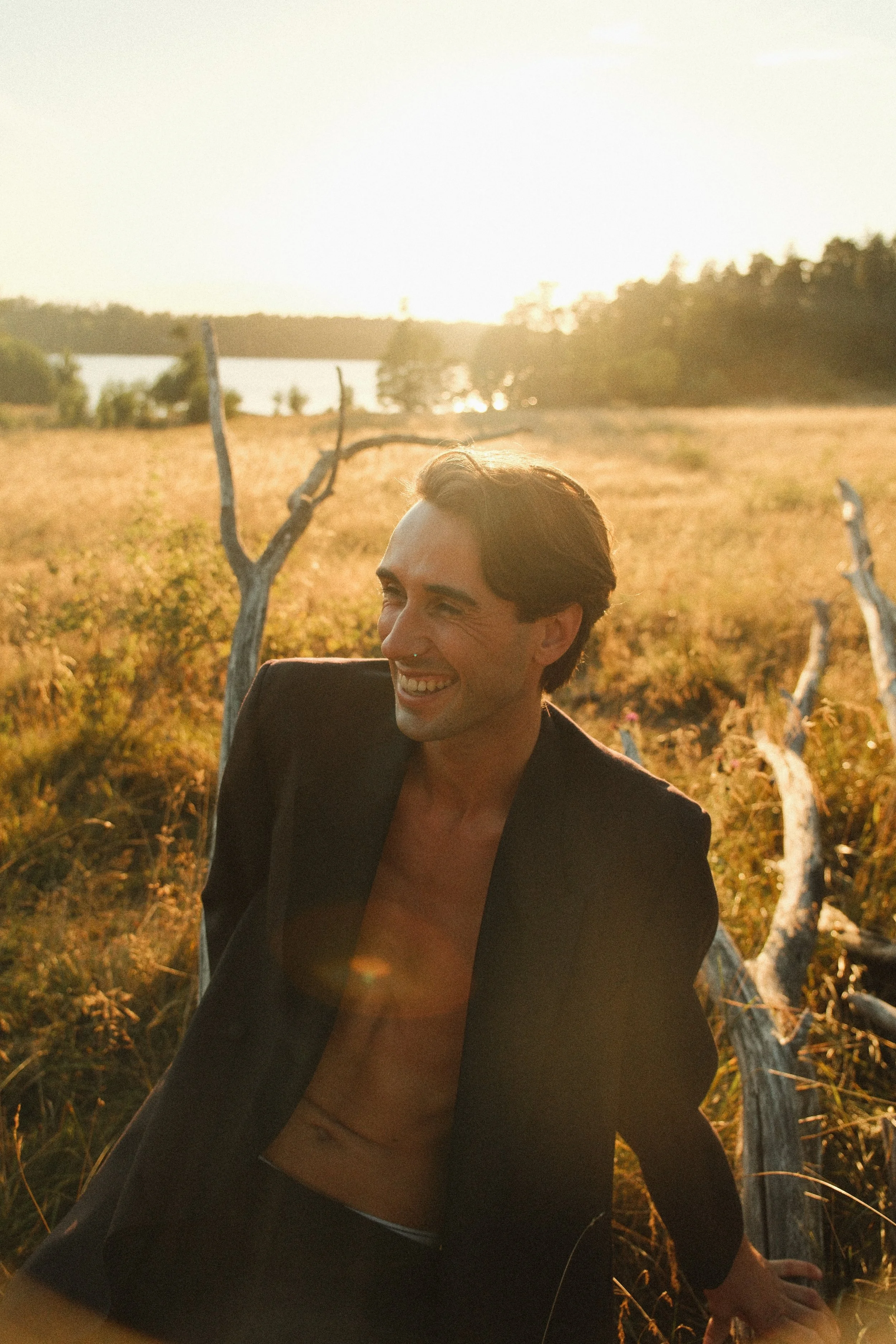 A woman smiling outdoors during sunset, sitting on a fallen tree trunk in a grassy field with trees and a body of water in the background.