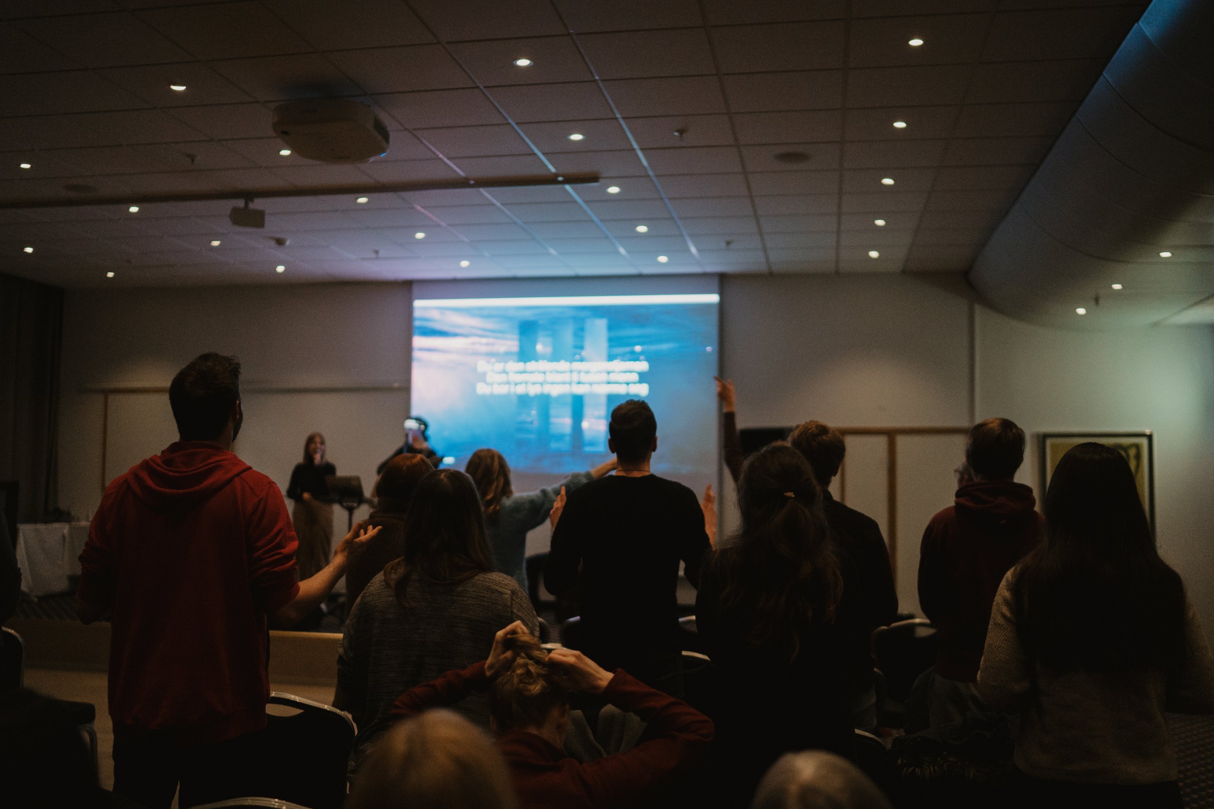 A group of people attending a presentation or seminar in a conference room with a large screen at the front displaying a slide. The attendees are facing the screen, and some are raising their hands.