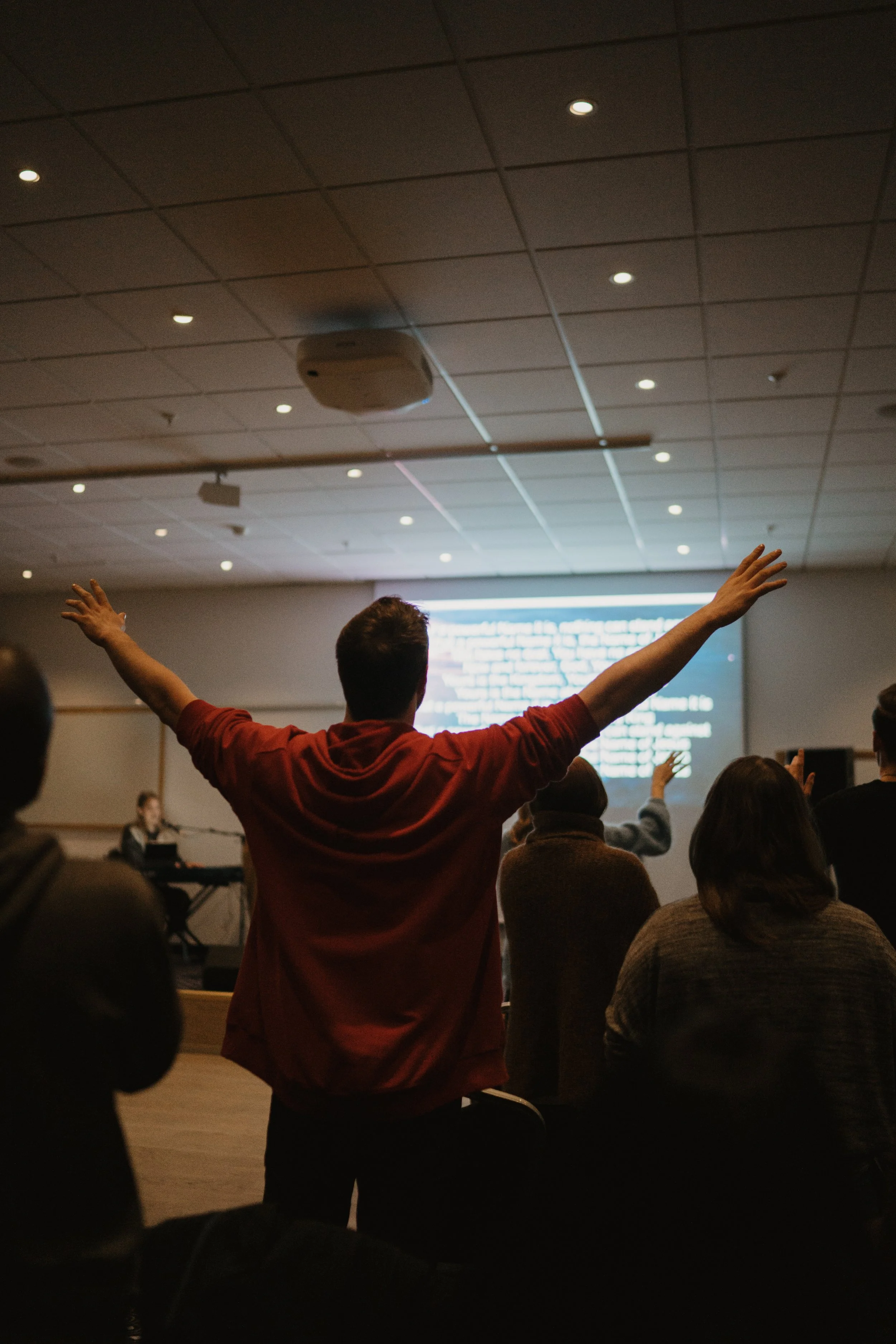 A man with arms raised stands in a dark room with an audience facing him. A large screen with text is visible in the background.