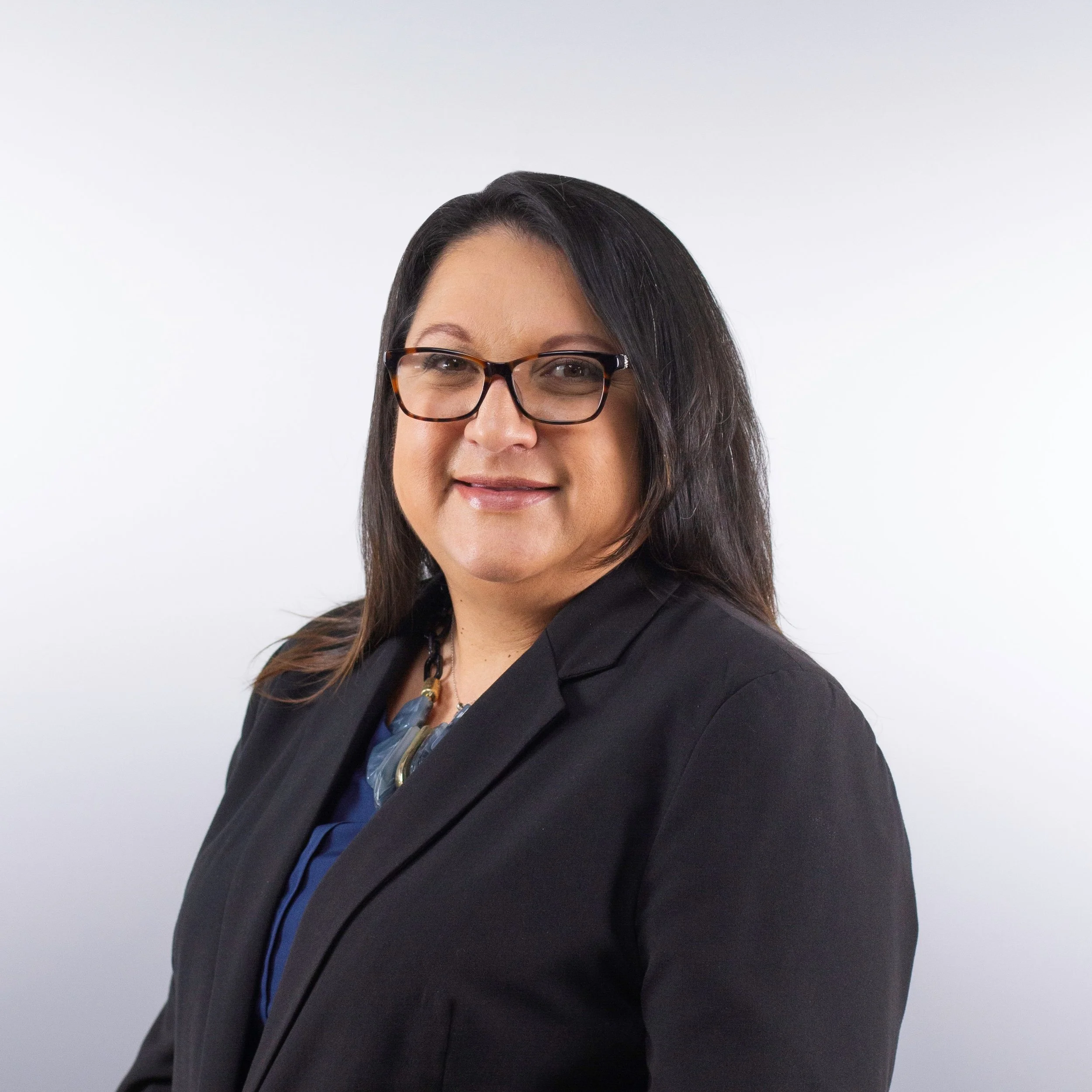 Close-up of a woman with black hair wearing glasses and a black blazer, smiling, against a plain white background.